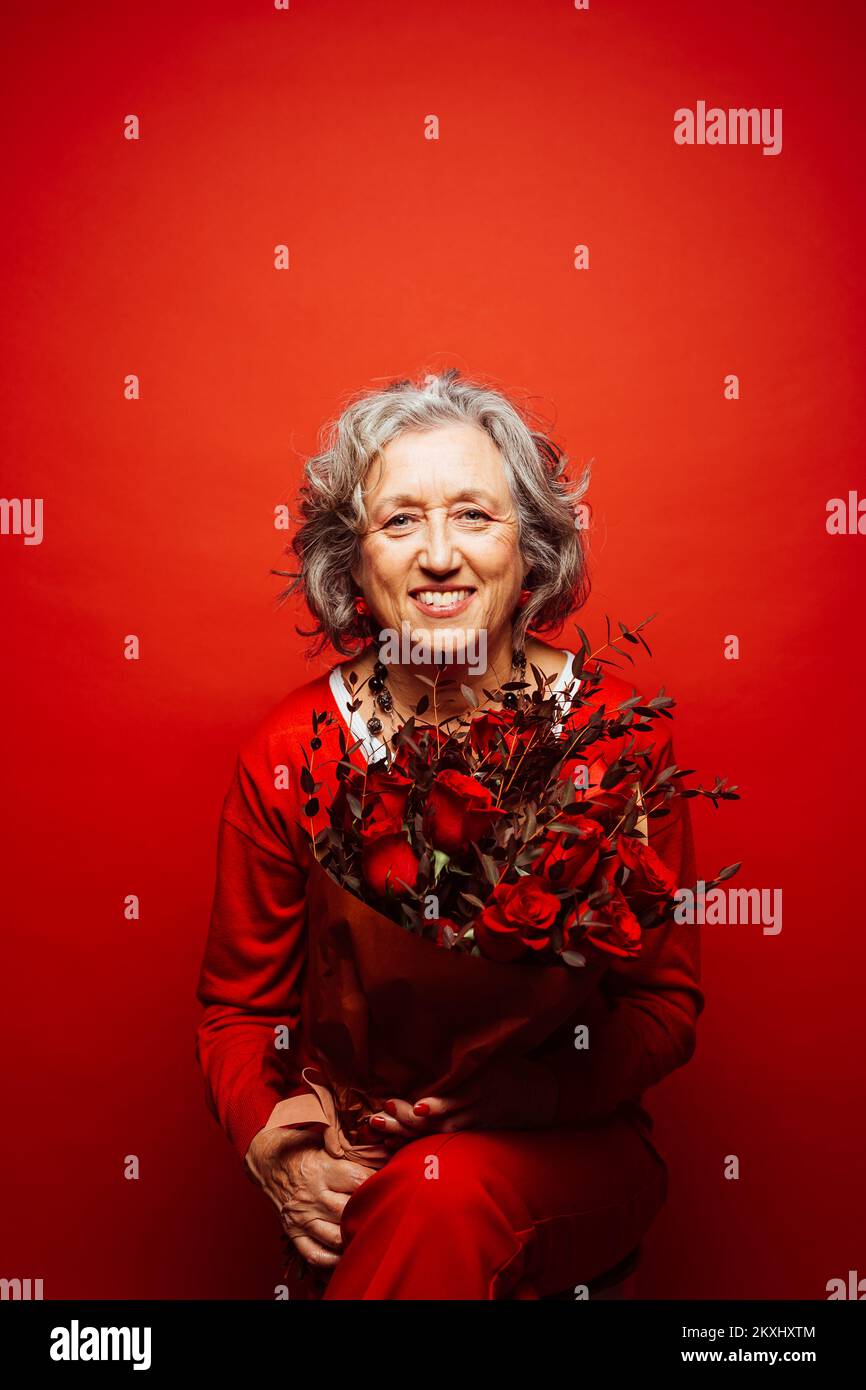Femme âgée portant des vêtements rouges, portant un bouquet de roses rouges, sur un fond rouge Banque D'Images