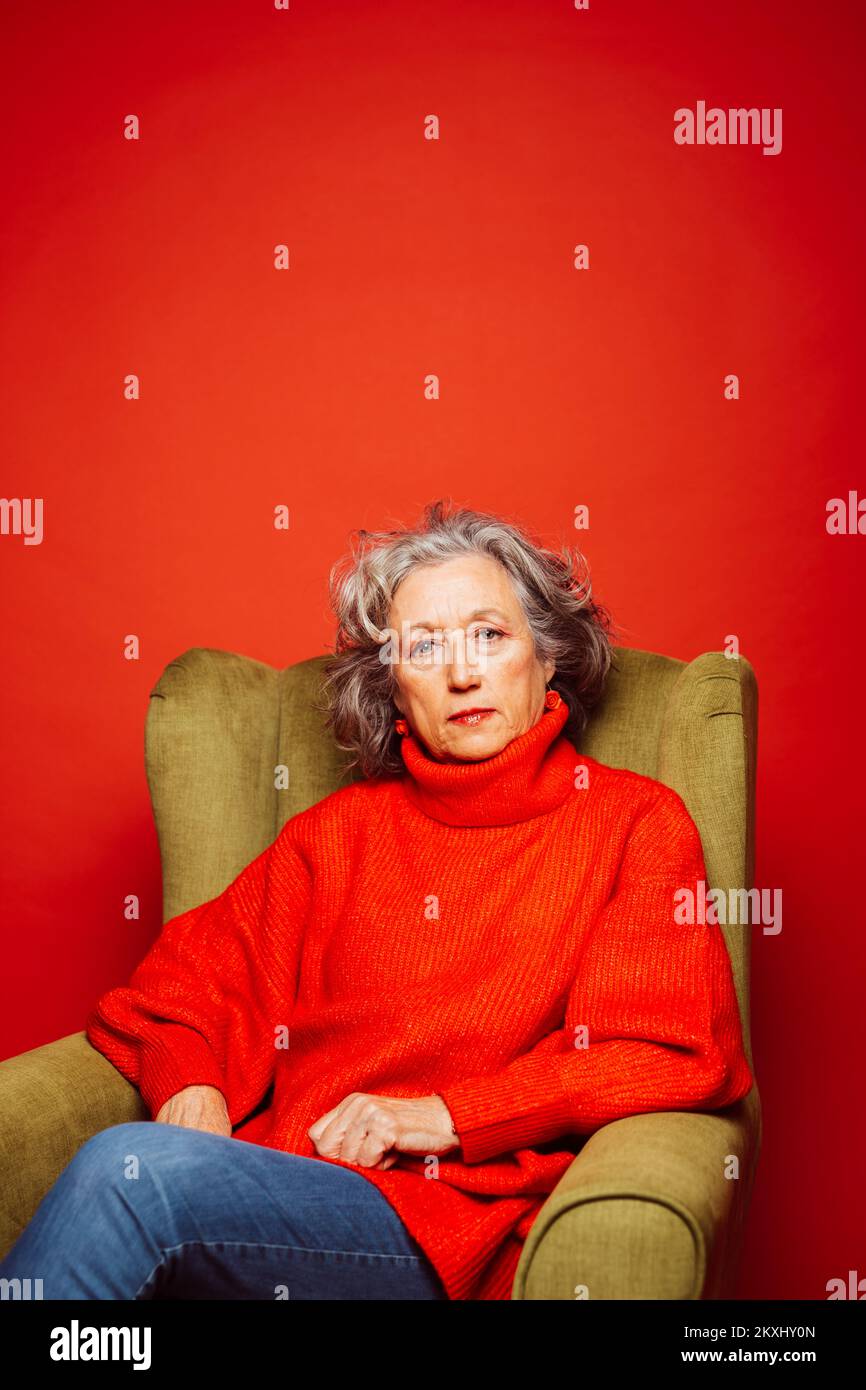 Femme âgée vêtue de vêtements rouges, assise sur un fauteuil vert, sur un fond rouge Banque D'Images Femme âgée vêtue de vêtements rouges, assise sur un fauteuil vert, sur un fond rouge Banque D'Images