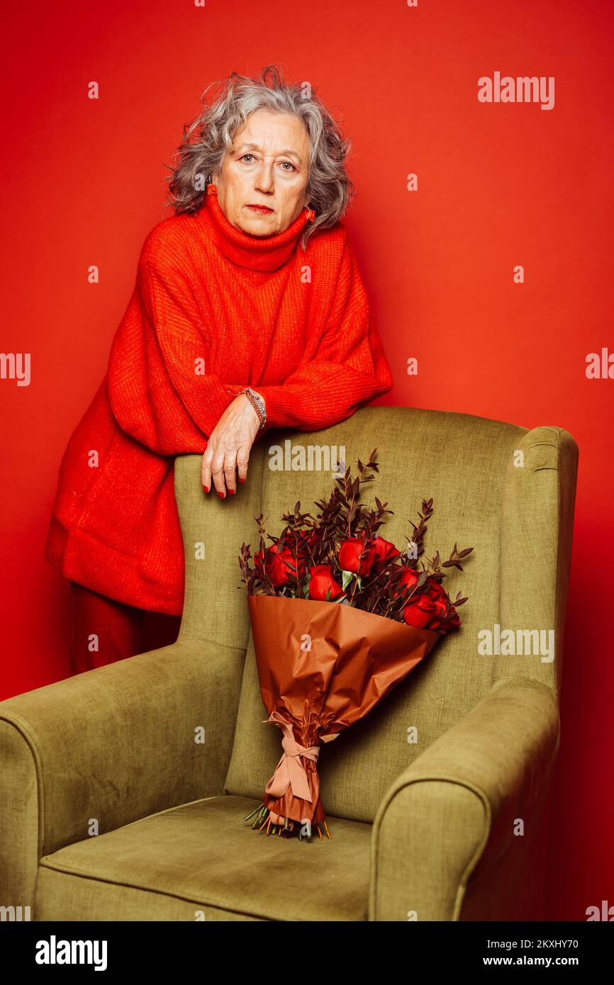 Femme âgée vêtue de vêtements rouges, debout à côté d'un fauteuil vert sur fond rouge Banque D'Images Femme âgée vêtue de vêtements rouges, debout à côté d'un fauteuil vert sur fond rouge Banque D'Images