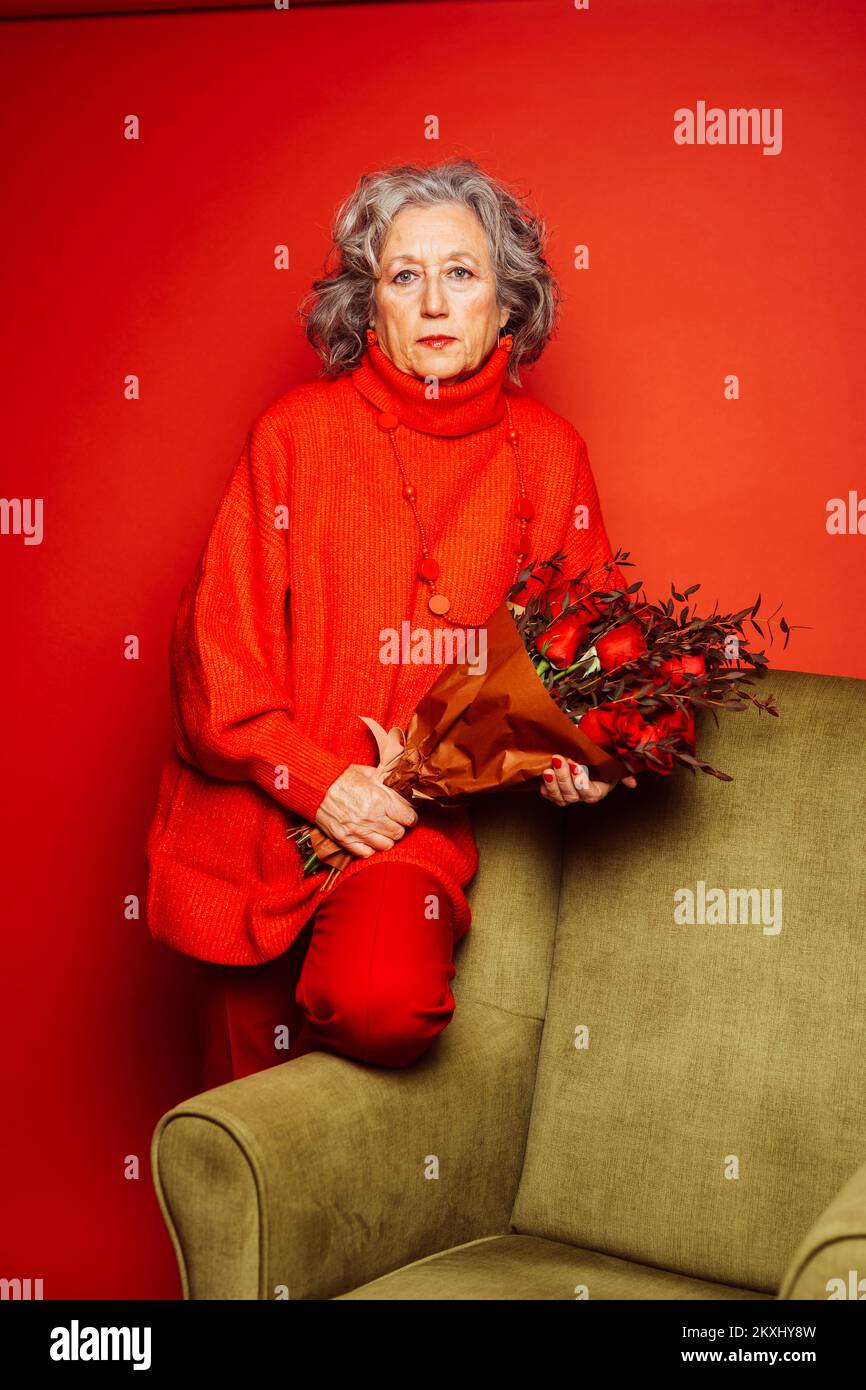 Femme âgée vêtue de vêtements rouges et assise sur un fauteuil vert avec un bouquet de roses rouges, sur un fond rouge Banque D'Images Femme âgée vêtue de vêtements rouges et assise sur un fauteuil vert avec un bouquet de roses rouges, sur un fond rouge Banque D'Images