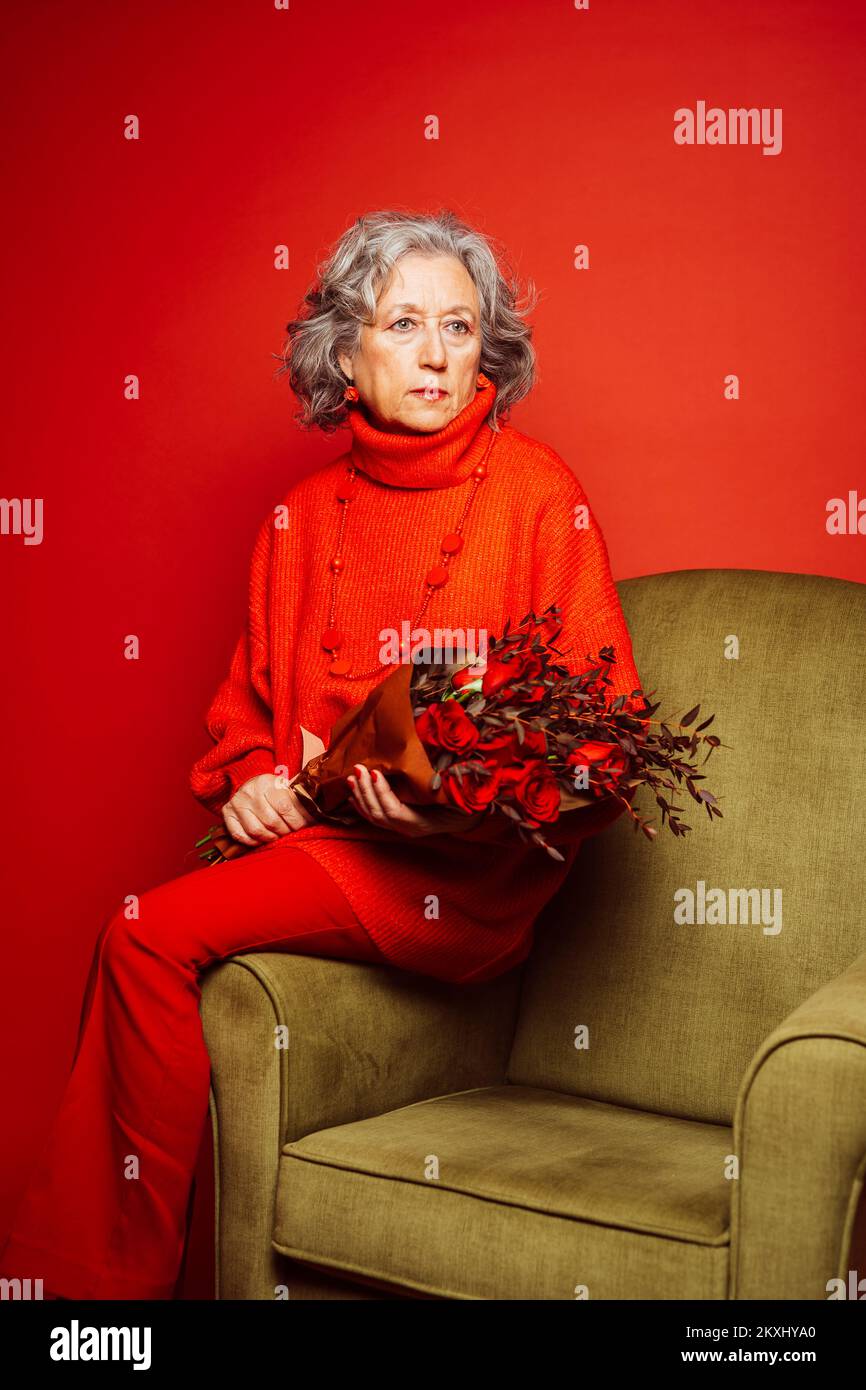 Femme âgée vêtue de vêtements rouges et assise sur un fauteuil vert avec un bouquet de roses rouges, sur un fond rouge Banque D'Images Femme âgée vêtue de vêtements rouges et assise sur un fauteuil vert avec un bouquet de roses rouges, sur un fond rouge Banque D'Images