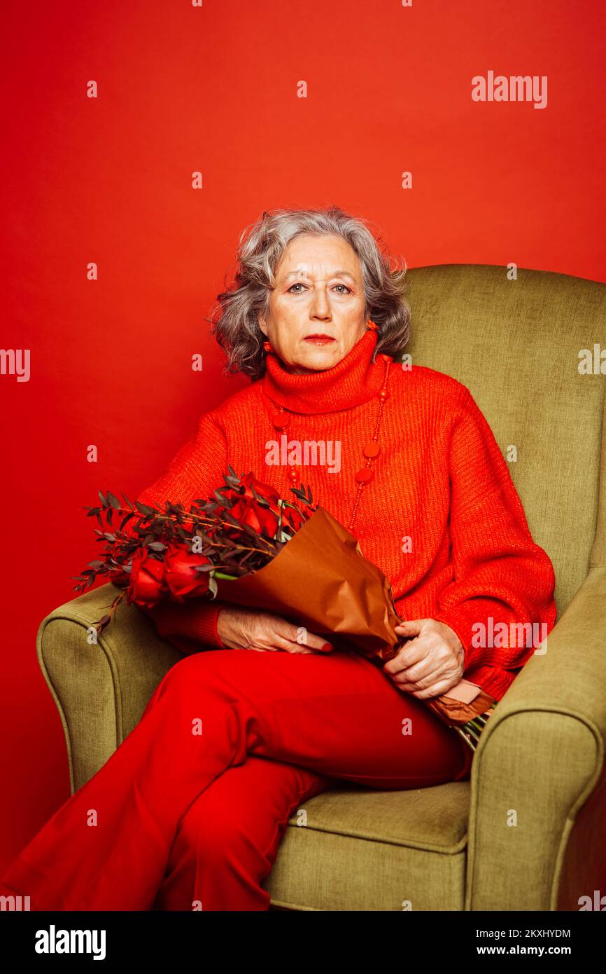 Femme âgée vêtue de vêtements rouges et assise sur un fauteuil vert avec un bouquet de roses rouges, sur un fond rouge Banque D'Images Femme âgée vêtue de vêtements rouges et assise sur un fauteuil vert avec un bouquet de roses rouges, sur un fond rouge Banque D'Images