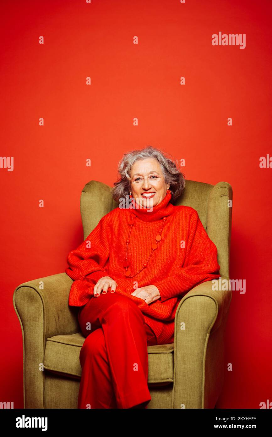 Femme âgée vêtue de vêtements rouges et assise sur un fauteuil vert sur fond rouge Banque D'Images Femme âgée vêtue de vêtements rouges et assise sur un fauteuil vert sur fond rouge Banque D'Images