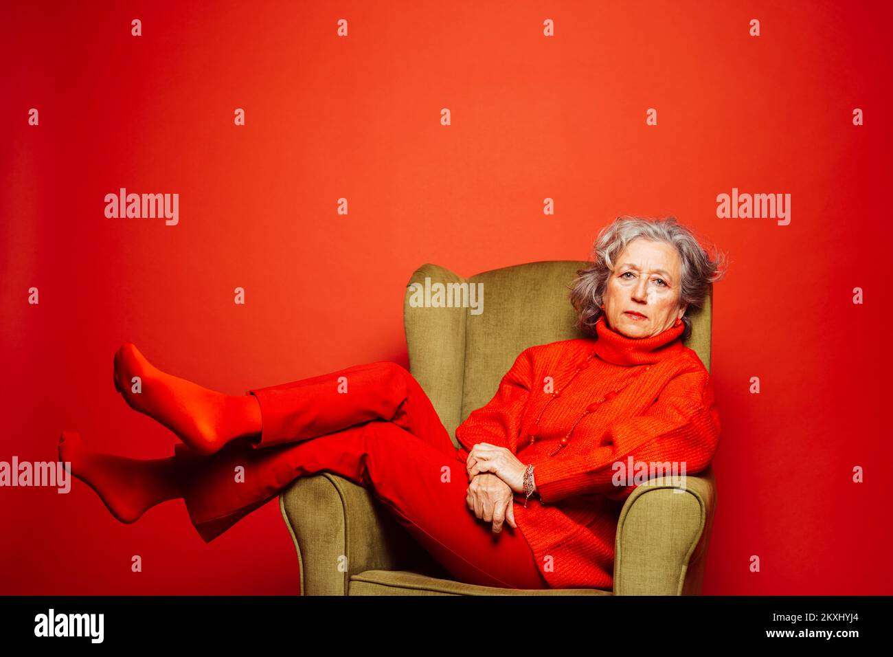 Femme âgée vêtue de vêtements rouges et assise sur un fauteuil vert sur fond rouge Banque D'Images Femme âgée vêtue de vêtements rouges et assise sur un fauteuil vert sur fond rouge Banque D'Images