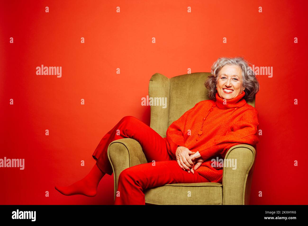 Femme âgée vêtue de vêtements rouges et assise sur un fauteuil vert sur fond rouge Banque D'Images Femme âgée vêtue de vêtements rouges et assise sur un fauteuil vert sur fond rouge Banque D'Images
