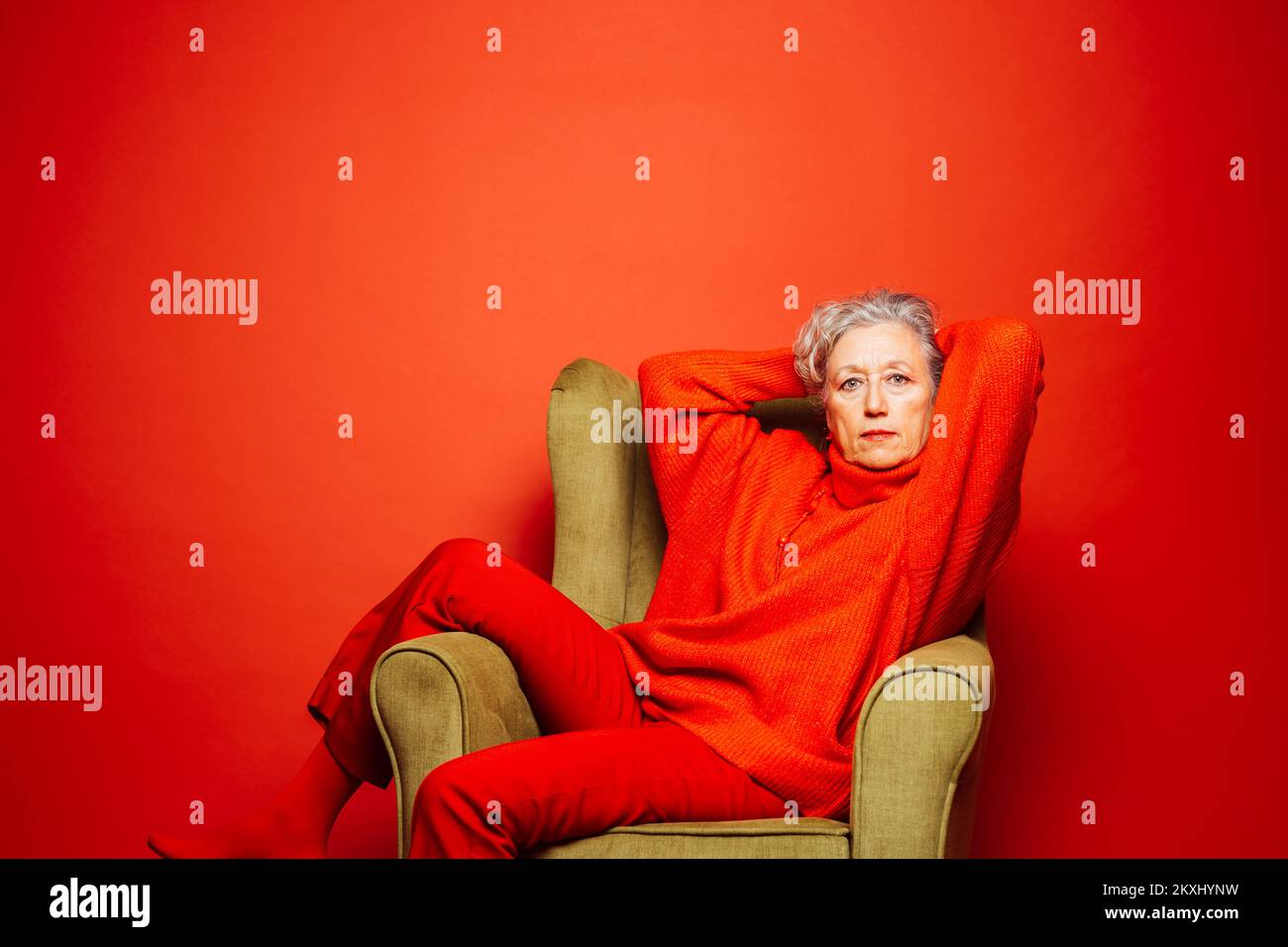 Femme âgée vêtue de vêtements rouges et assise sur un fauteuil vert sur fond rouge Banque D'Images