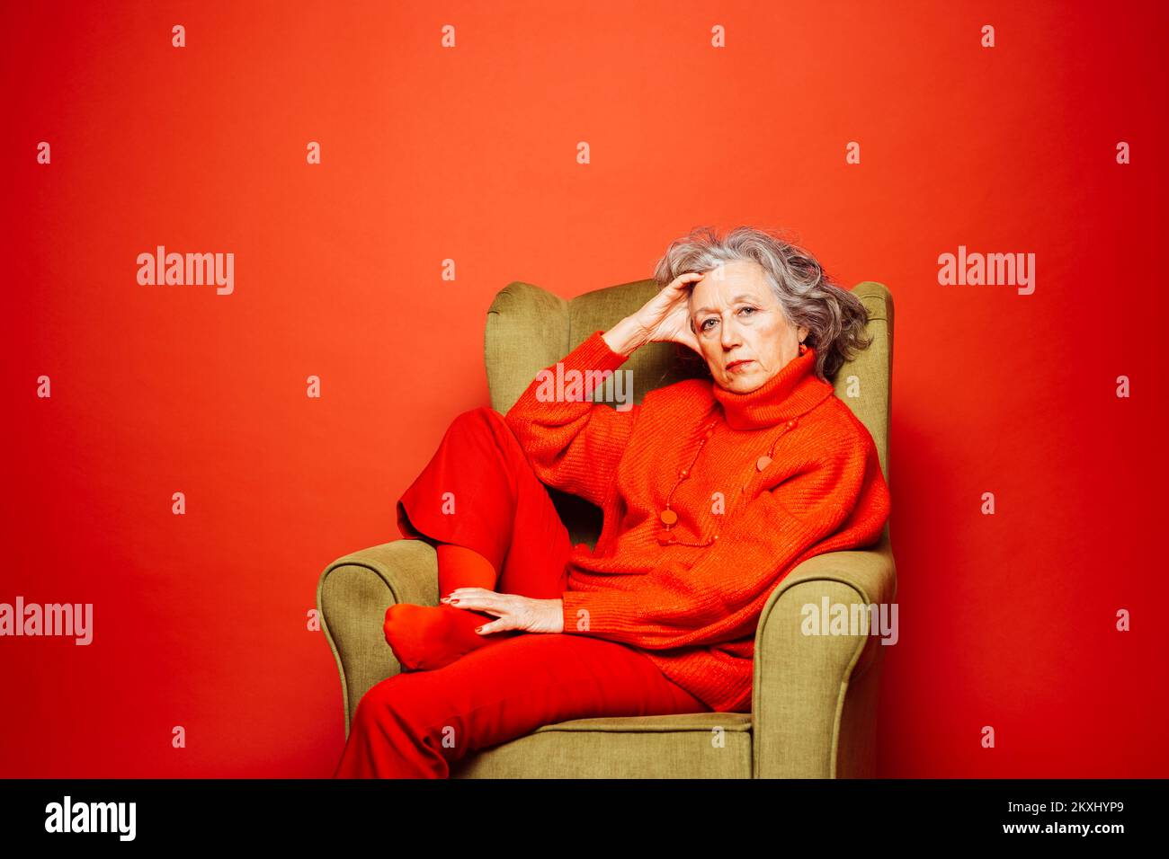 Femme âgée vêtue de vêtements rouges et assise sur un fauteuil vert sur fond rouge Banque D'Images Femme âgée vêtue de vêtements rouges et assise sur un fauteuil vert sur fond rouge Banque D'Images