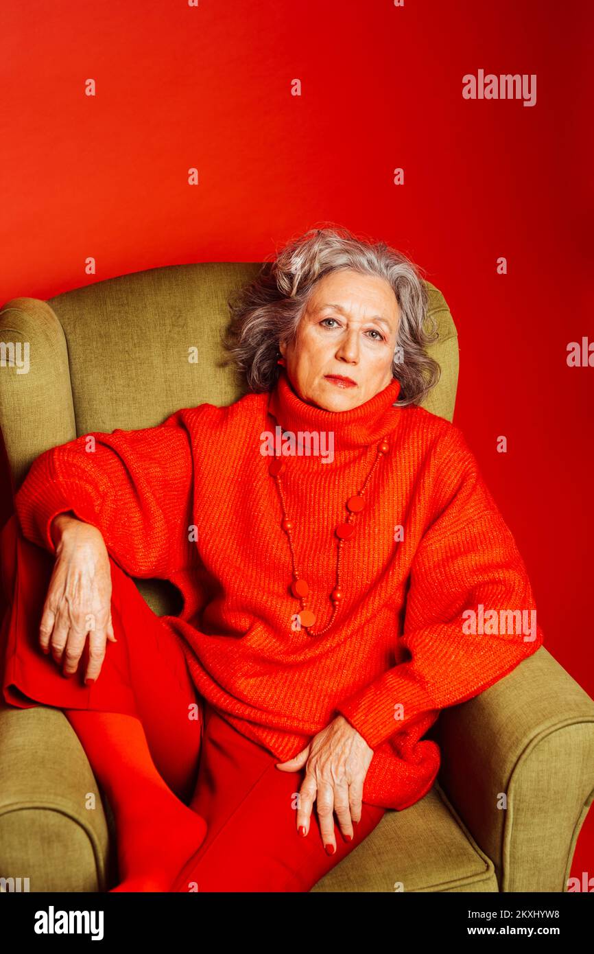 Femme âgée vêtue de vêtements rouges et assise sur un fauteuil vert sur fond rouge Banque D'Images Femme âgée vêtue de vêtements rouges et assise sur un fauteuil vert sur fond rouge Banque D'Images