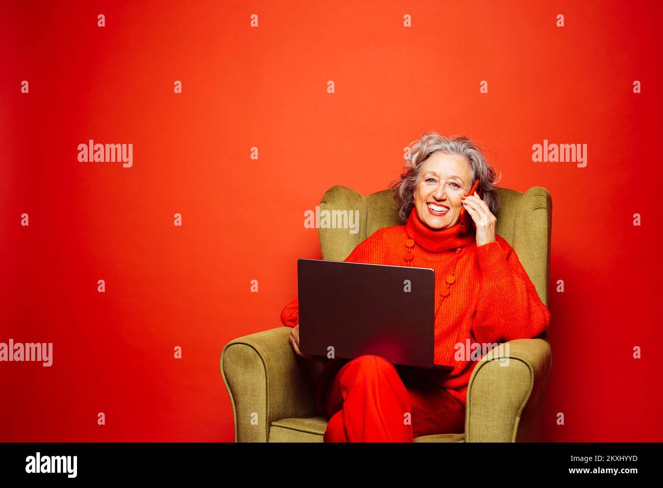 Femme âgée, vêtue de vêtements rouges et assise sur un fauteuil vert, utilisant un ordinateur portable et un téléphone sur un fond rouge Banque D'Images