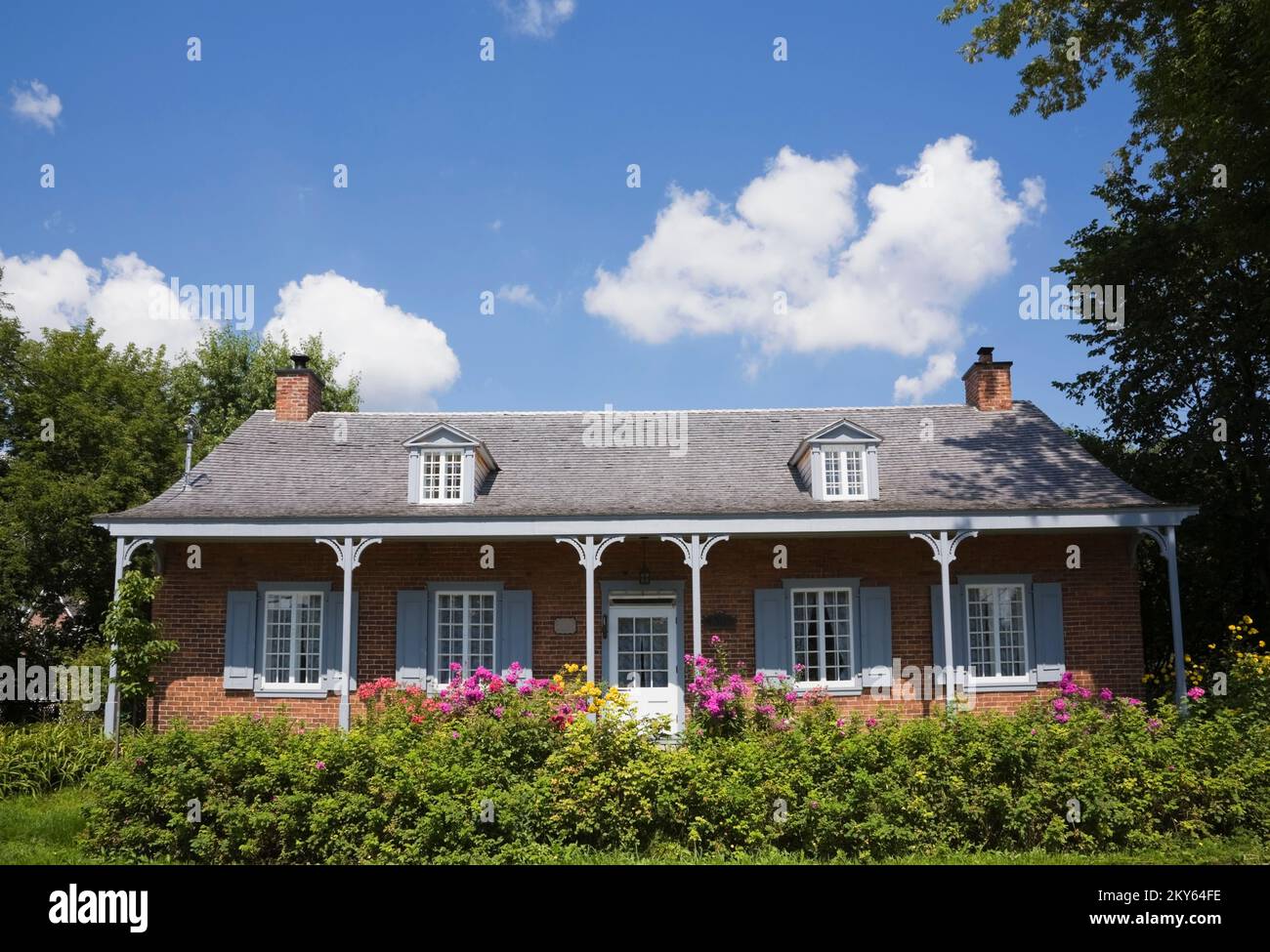 Maison ancienne en briques nuancées rouges 1858 avec bordure grise et blanche. grande véranda et cour avant paysagée en été. Banque D'Images