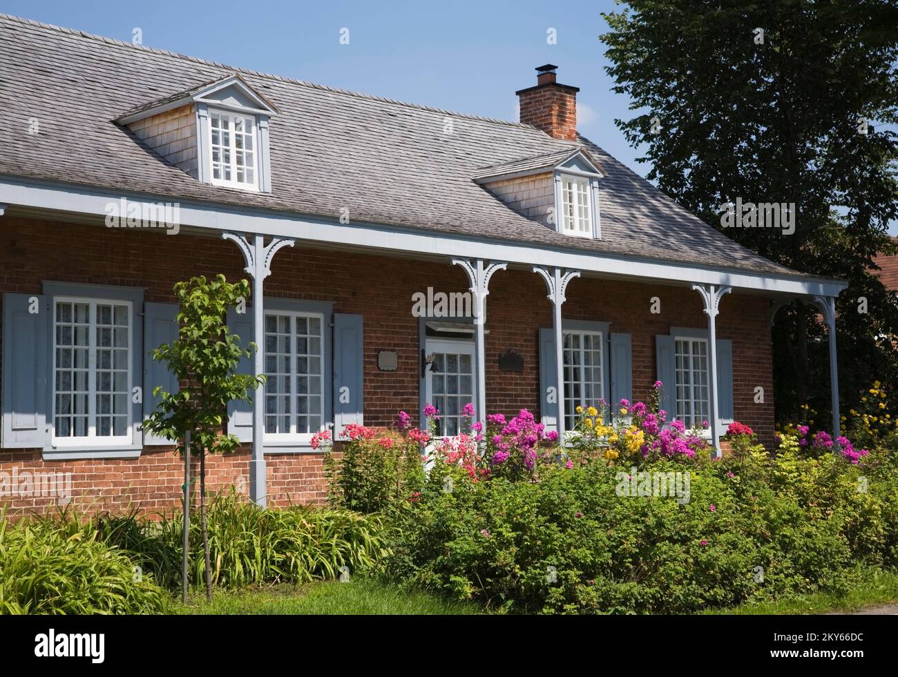 Maison ancienne en briques nuancées rouges 1858 avec bordure grise et blanche. grande véranda et cour avant paysagée en été. Banque D'Images