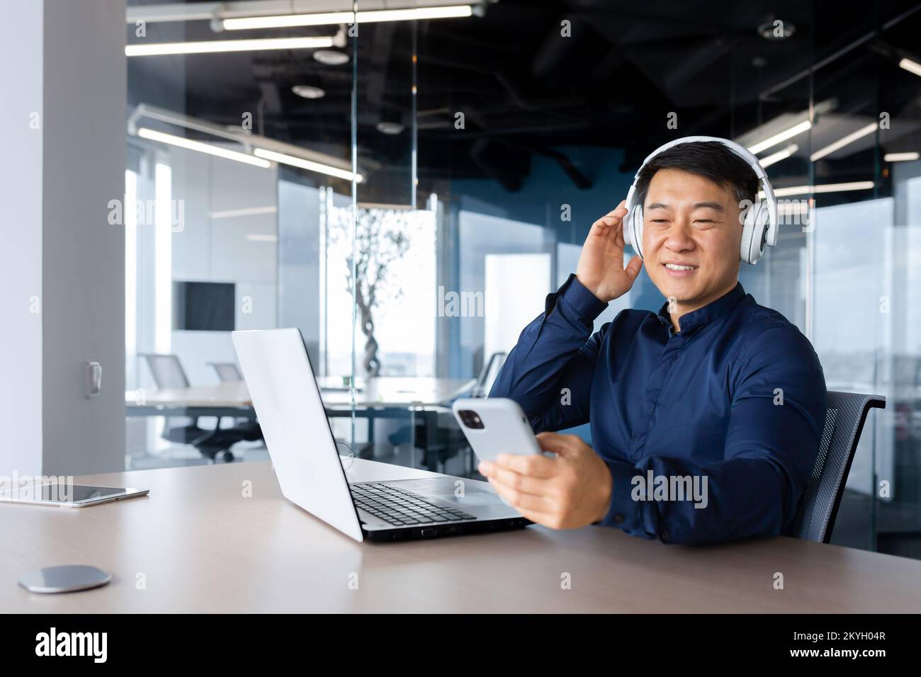 Homme d'affaires à l'écoute de musique et de podcast sur le lieu de travail, homme asiatique souriant et heureux d'utiliser un ordinateur portable au travail, homme avec casque et téléphone à l'écoute de la radio en ligne dans le bureau. Banque D'Images
