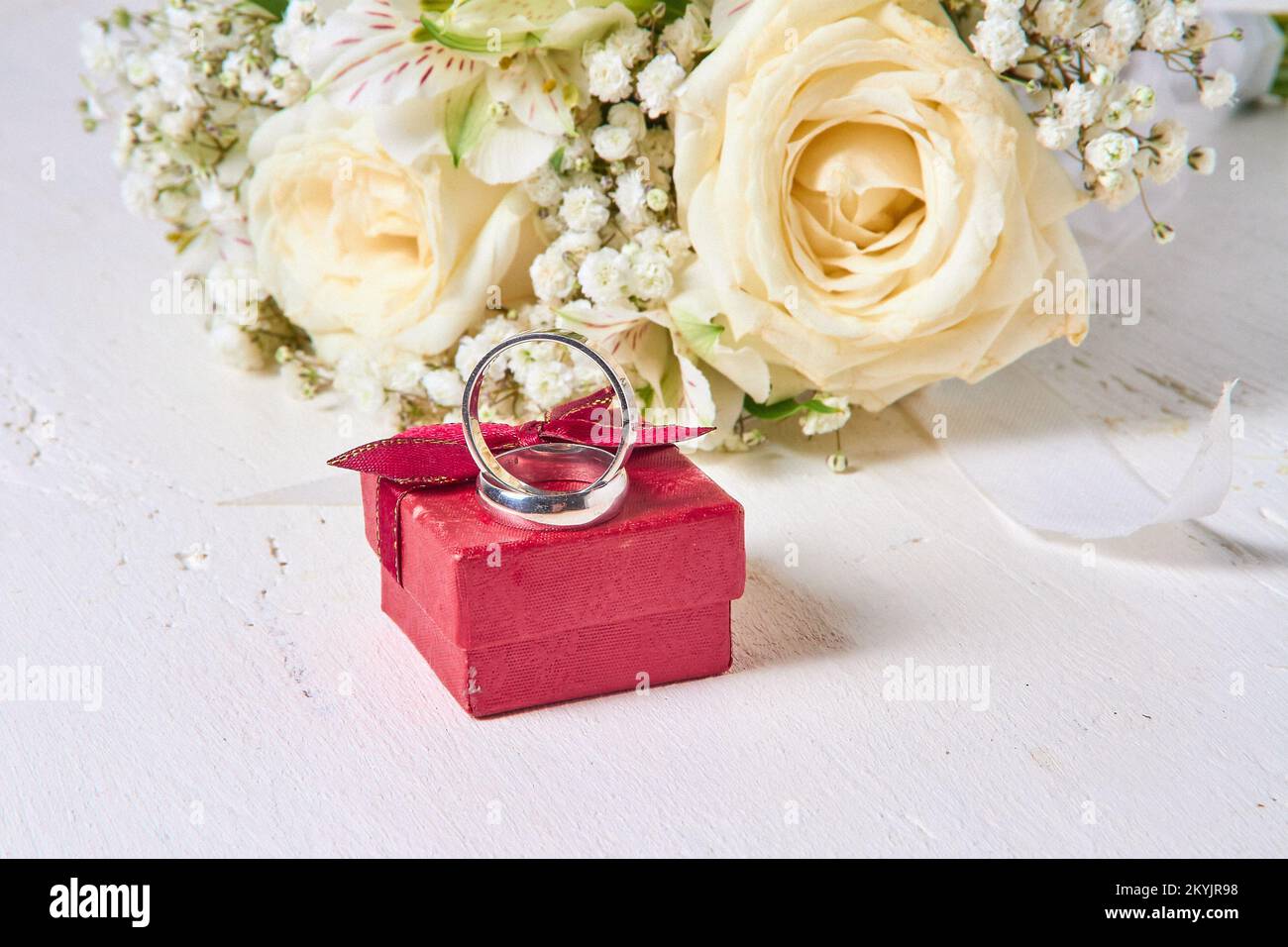 Deux anneaux de mariage de différentes tailles, une boîte cadeau rouge et un bouquet de mariée se trouvent sur un fond blanc en bois. Banque D'Images