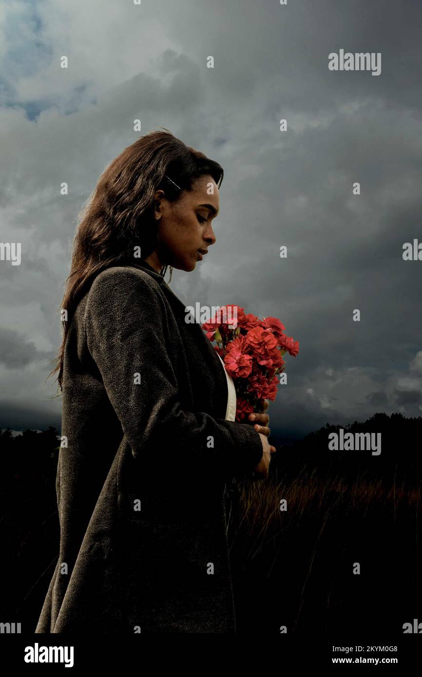 jeune femme noire tenant un bouquet de fleurs rouges regardant la mélancolie. porter une chemise blanche et un manteau foncé. Banque D'Images
