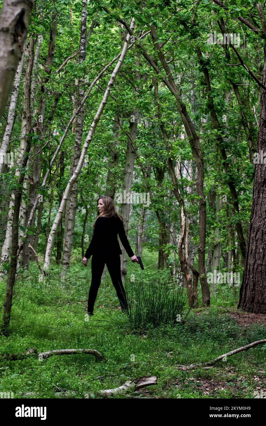 Jeune femme aux cheveux rouges vêtue de noir et d'un pistolet dans les bois, thriller, mystère, style couverture de livre d'aventure. Banque D'Images