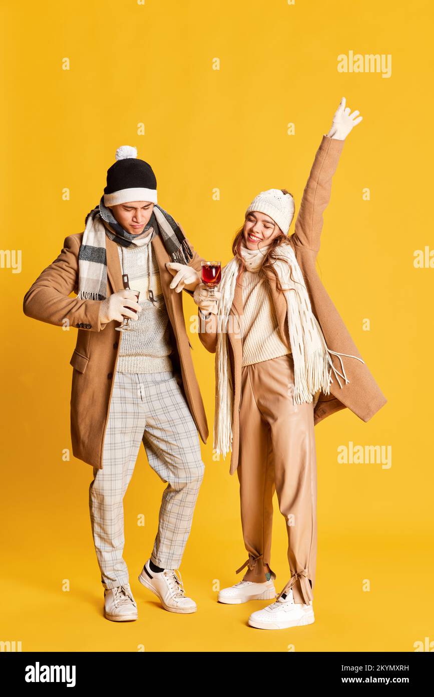 Portrait de jeune homme et femme élégant et gai dans des vêtements d'hiver confortables posant, buvant du vin chaud isolé sur fond jaune Banque D'Images