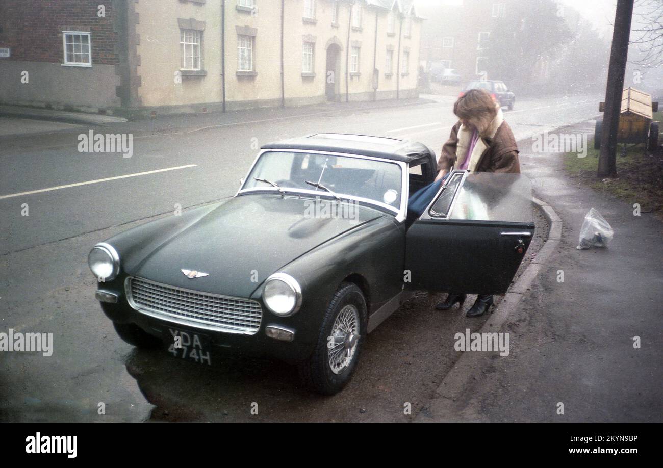 1980s, une séance photo tôt le matin et une jeune femme portant une veste en cuir luttant pour obtenir des sacs d'équipements de caméra dans une petite voiture de sport Austin-Healey Sprite de 2 portes, garée dans un laprès d'une route, Angleterre, Royaume-Uni. Un sac avec une lentille d'appareil photo se trouve sur la chaussée. En production de 1958 à 1971, la Sprite a été conçue par la Donald Healey Motor Company et fabriquée à l'usine MG d'Abingdon, à Oxford, en Angleterre. MG avait sa propre petite voiture de sport 2 portes, le Midget, qui était pratiquement identique à l'Austin-Healey Sprite mais avec un badge différent. Banque D'Images