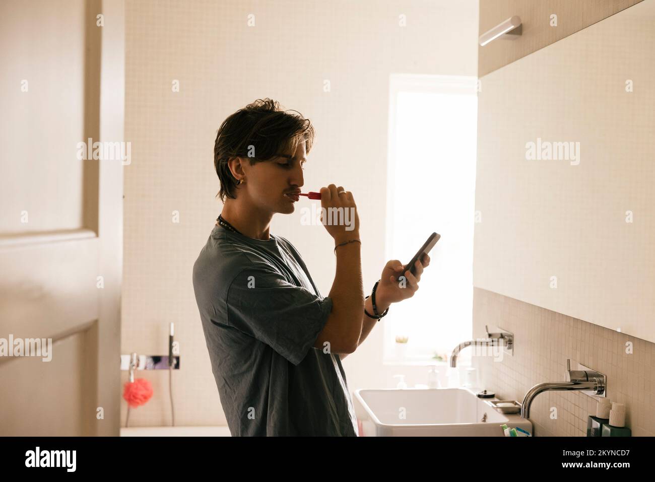 Vue latérale d'un jeune homme utilisant un smartphone tout en se brossant les dents dans la salle de bains à la maison Banque D'Images