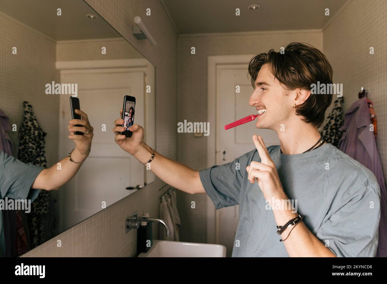 Homme souriant prenant le selfie à travers le smartphone tout en se brossant les dents dans la salle de bains à la maison Banque D'Images