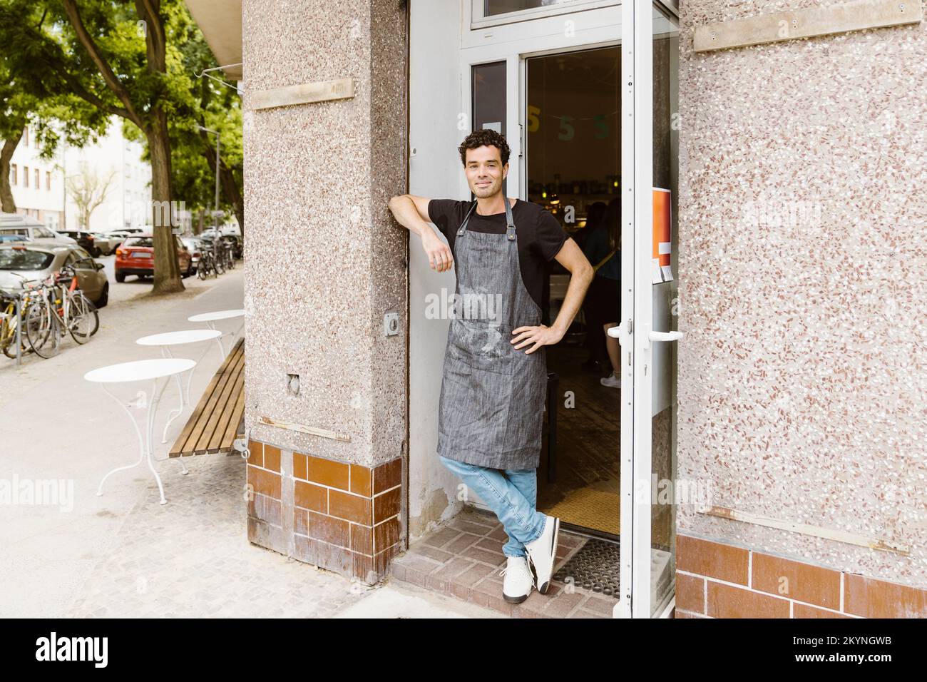 Un homme souriant propriétaire de café avec la main sur la hanche penchée sur le mur à la porte Banque D'Images