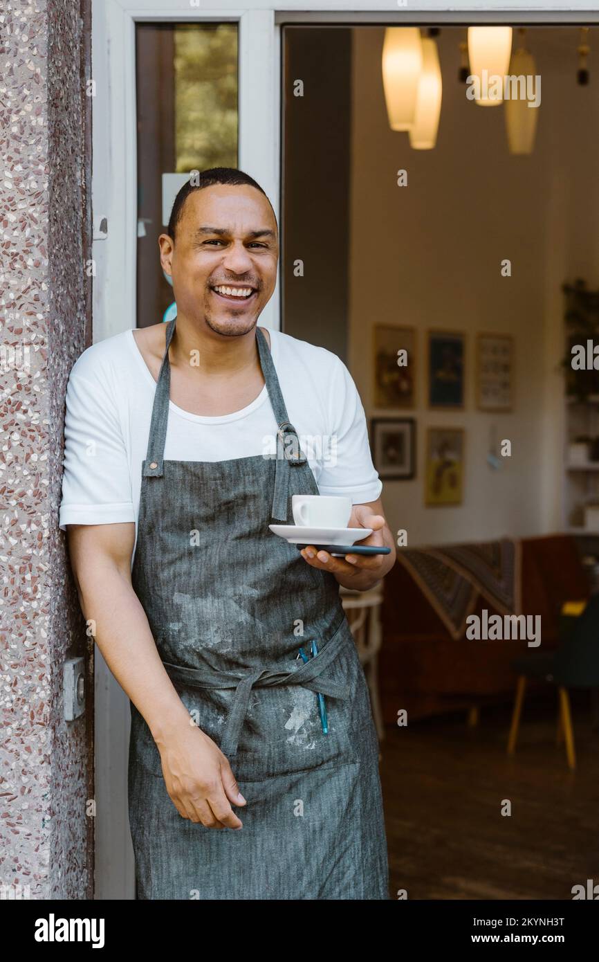 Portrait d'un homme heureux portant un tablier tenant une tasse de café tout en se penchant à la porte du café Banque D'Images
