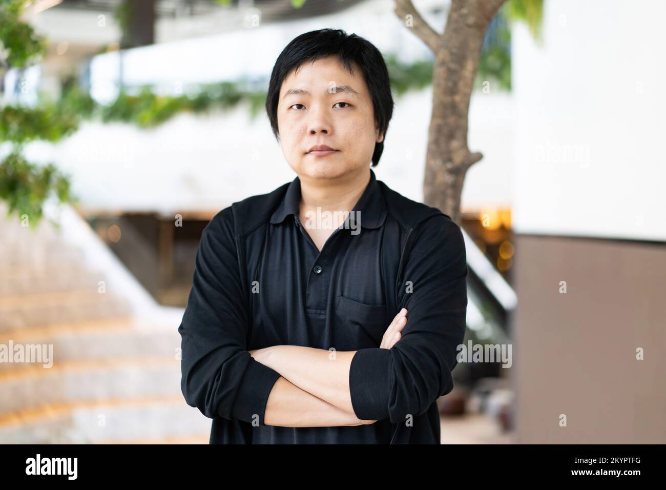 Le portrait d'un homme asiatique dans une chemise noire décontractée avec des bras croisés debout devant un arrière-plan flou d'un espace de travail de bureau. Banque D'Images