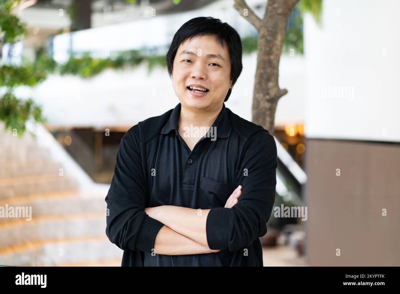 Le portrait d'un homme asiatique dans une chemise noire décontractée avec des bras croisés debout devant un arrière-plan flou d'un espace de travail de bureau. Banque D'Images