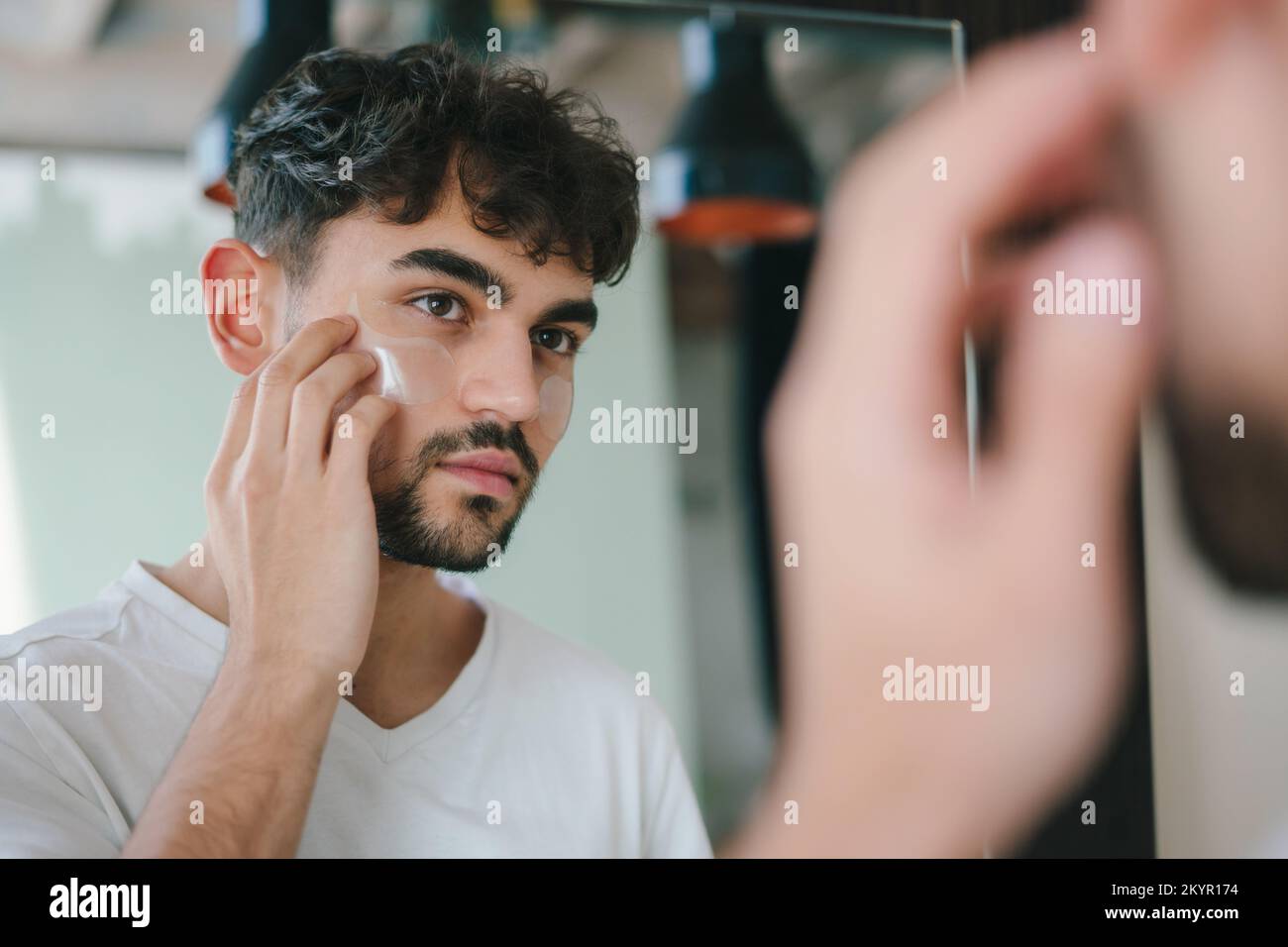 Homme barbu appliquant des taches oculaires sous ses yeux tout en regardant le miroir. Rides et soin du visage à domicile pour les hommes. Soin de la peau. Concept de soins à domicile Banque D'Images