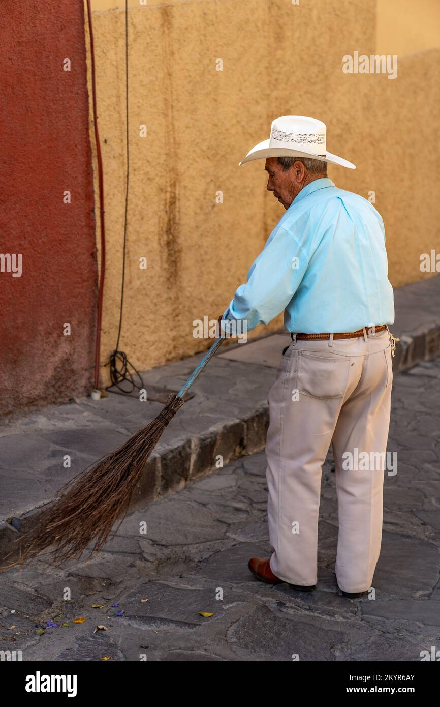 Un homme local portant un chapeau de paille et tenant un balai fait main balaie la rue devant sa maison à San Miguel de Allende, Mexique Banque D'Images