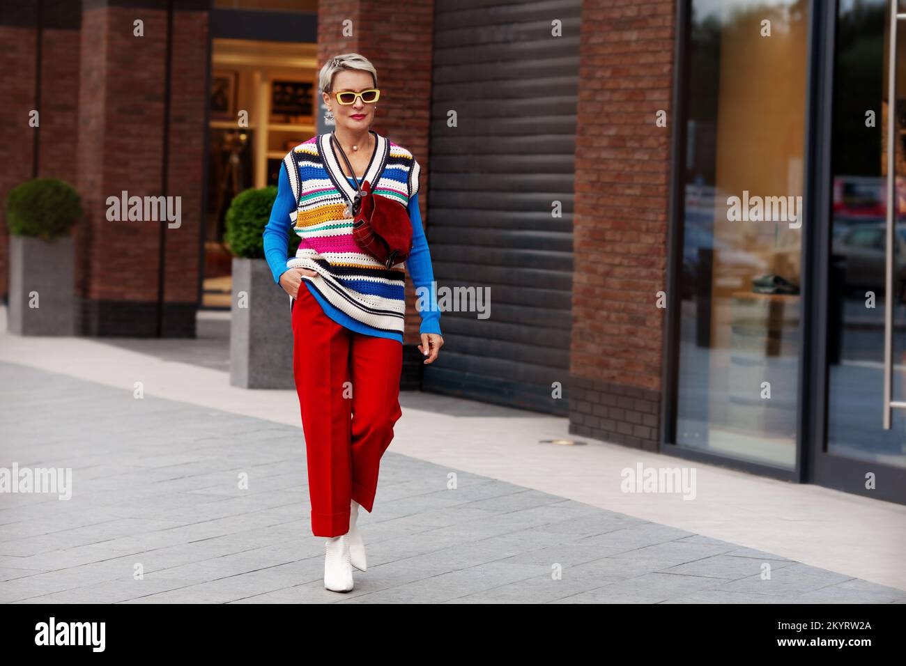Femme tendance portant une tenue tendance, un pantalon rouge, une veste bleue, un gilet rayé, des chaussures blanches et des lunettes de soleil jaunes. Modèle avec cheveux courts marchant le Banque D'Images
