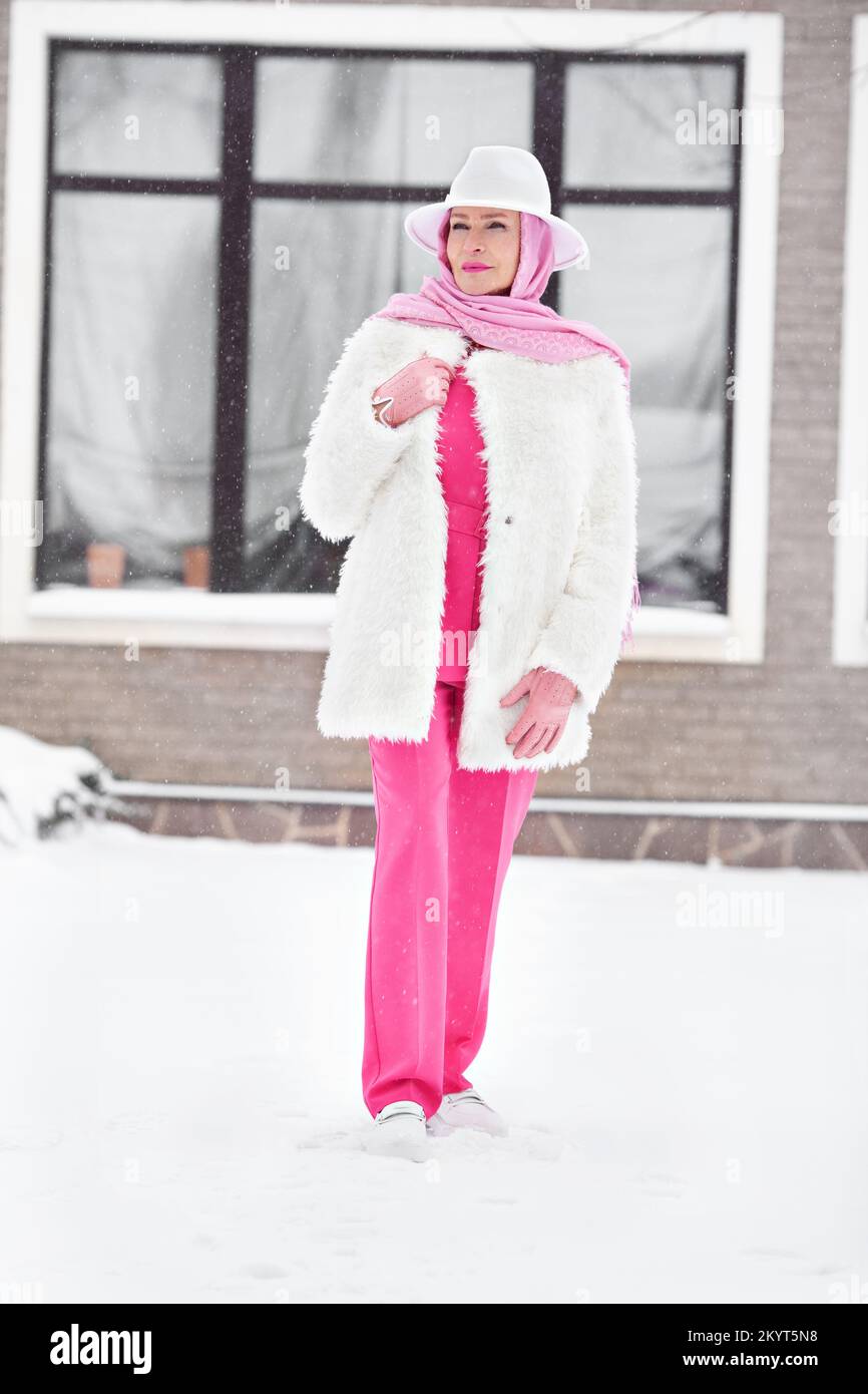 Femme mûre tendance en costume rose avec pantalon, foulard, chapeau en feutre blanc, manteau en fausse fourrure, tenue de style rue d'hiver Banque D'Images