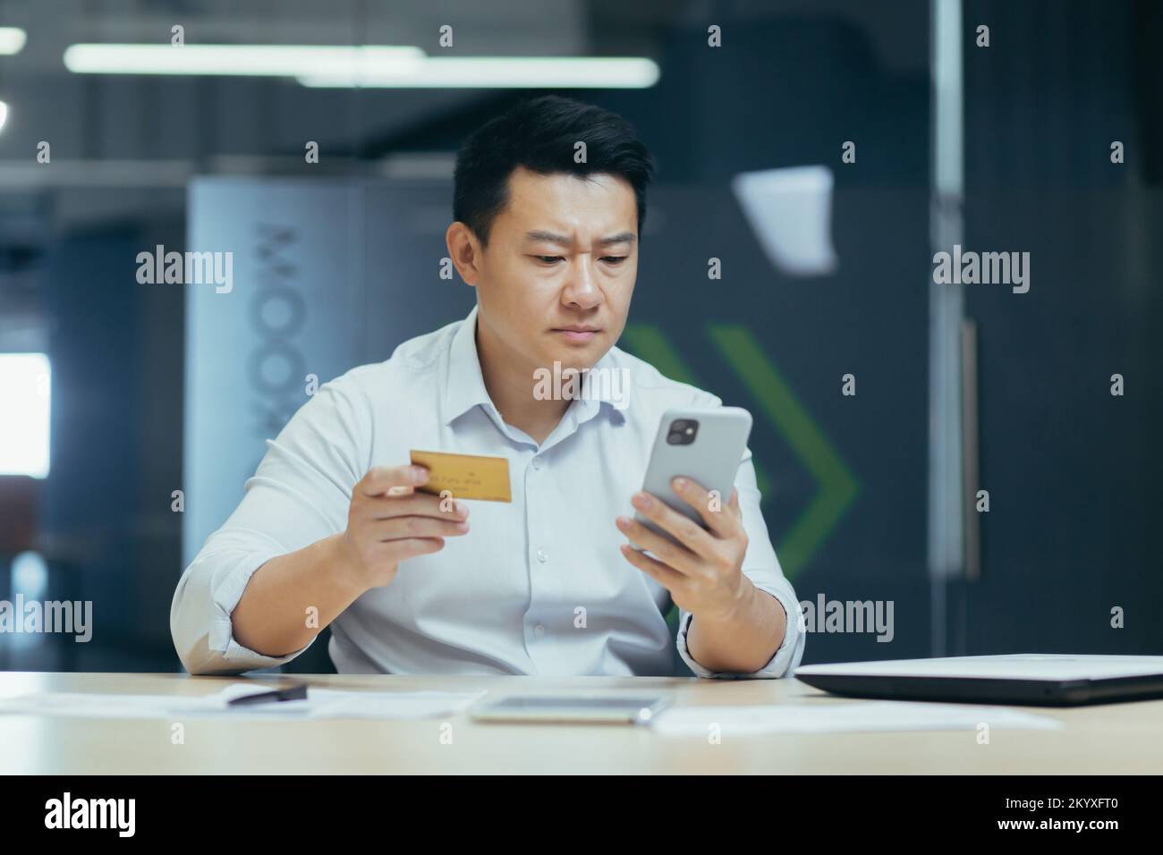Un jeune homme asiatique sérieux tient une carte de crédit et un téléphone. Effectue des transactions monétaires, des achats en ligne, vérifie le compte. Assis au bureau. Banque D'Images