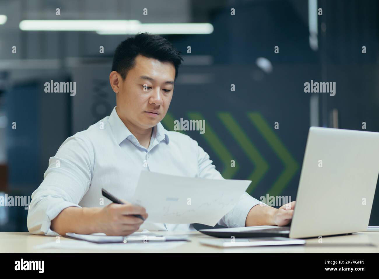Un jeune homme sérieux, un avocat asiatique, est assis dans le bureau du bureau, tient des documents et des accords entre ses mains. Travail concentré sur un ordinateur portable. Banque D'Images