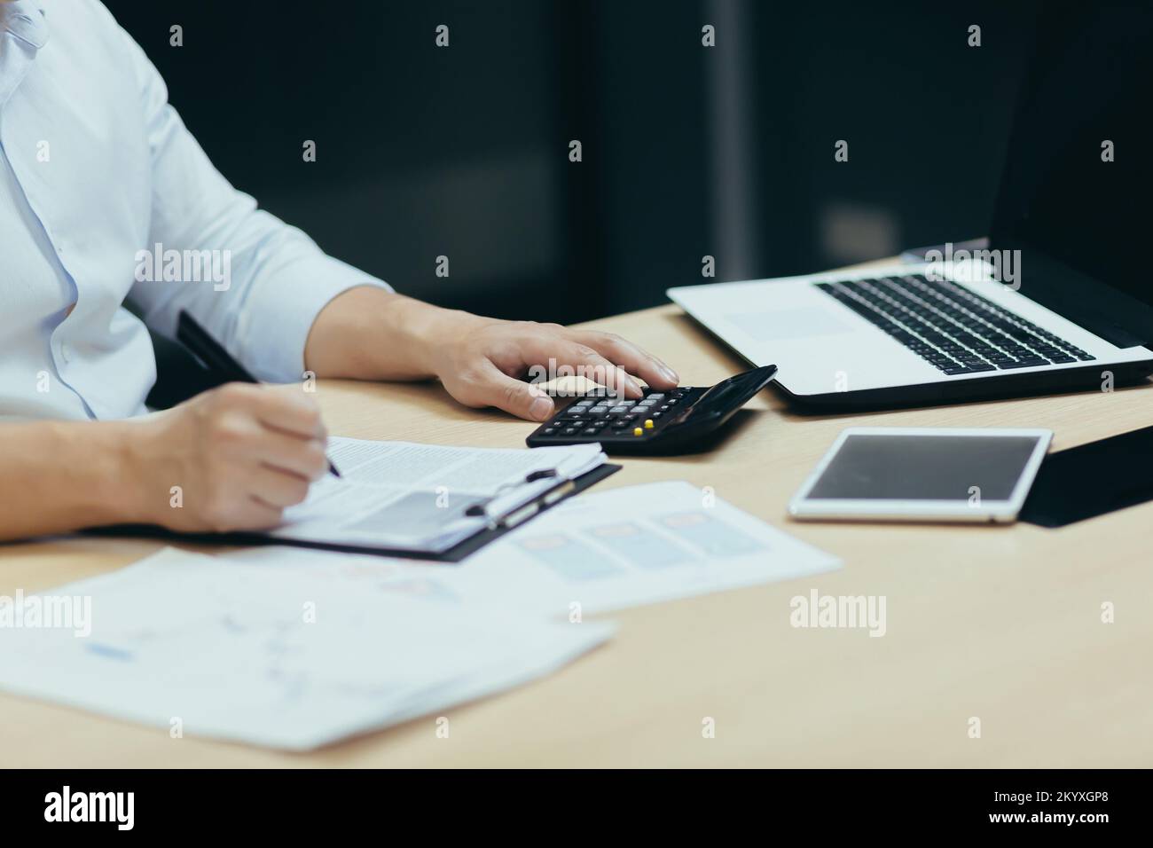 Gros plan. Les mains des hommes dans une chemise blanche à la table au bureau avec une tablette, un ordinateur portable, une calculatrice. Ils écrivent avec un stylo sur papier, signent des documents, travaillent avec des comptes et des finances, des rapports Banque D'Images