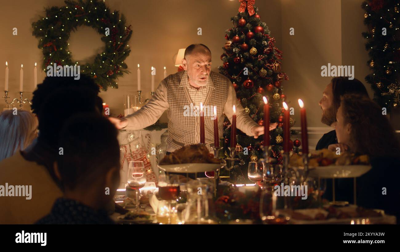 Grand-père âgé raconte une blague ou une histoire de vie à sa grande famille multiculturelle. Ils célèbrent Noël. Table avec plats et bougies. Ambiance chaleureuse du dîner de Noël familial à la maison. Banque D'Images