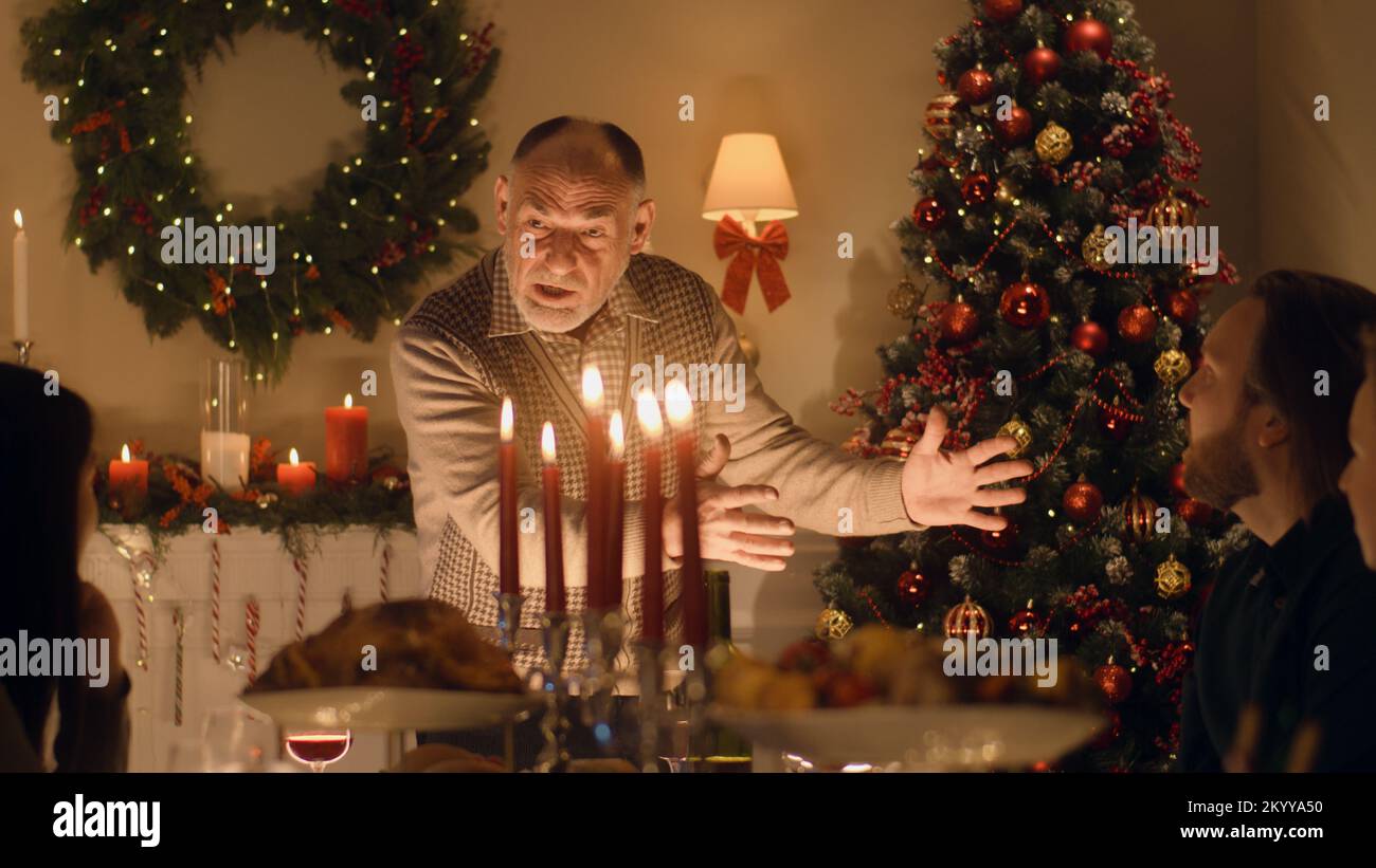 Un homme âgé raconte une blague ou une histoire de vie à sa grande famille multiculturelle. Ils célèbrent Noël. Table avec plats et bougies. Ambiance chaleureuse du dîner de Noël familial à la maison. Banque D'Images