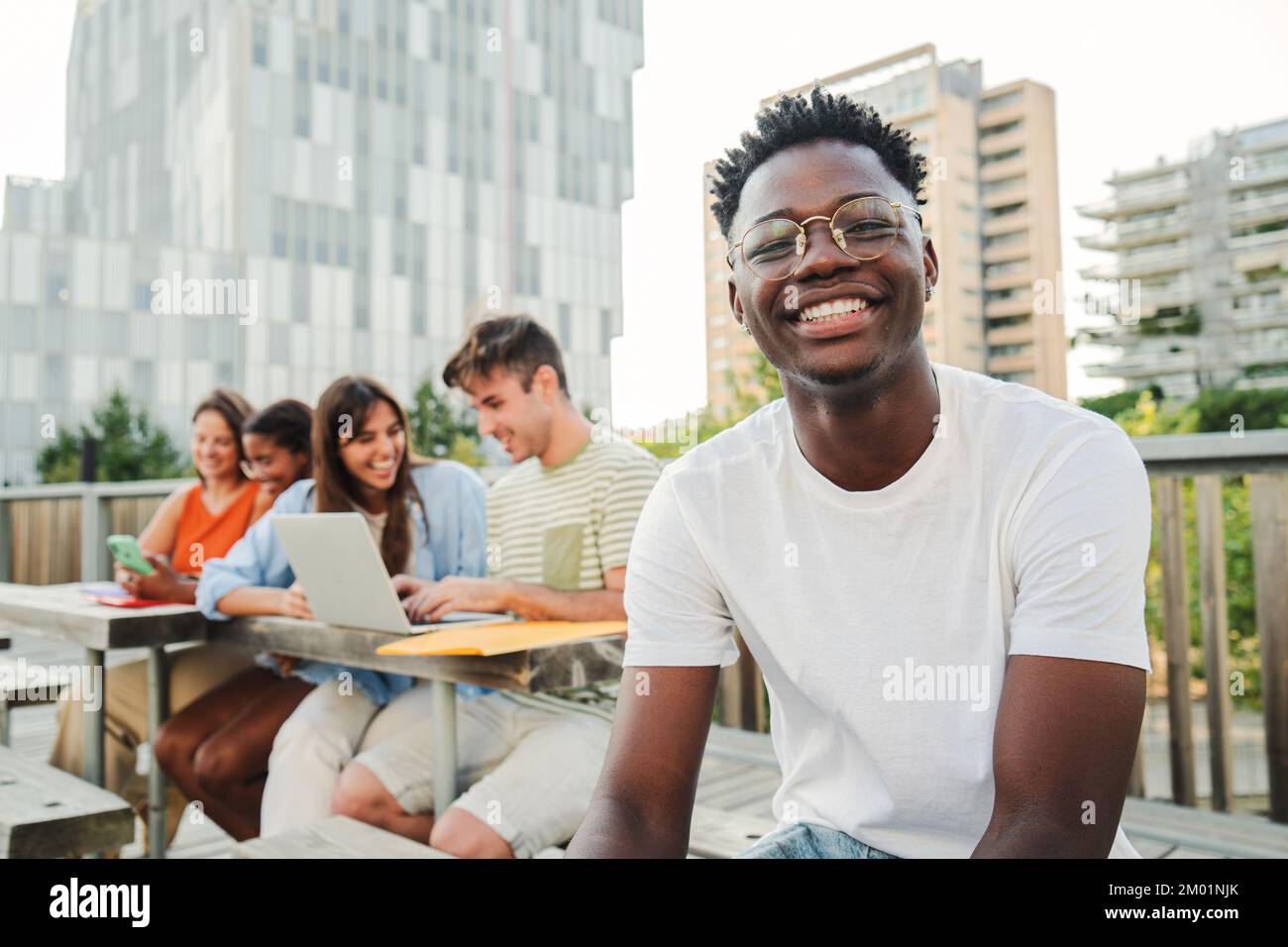 Au premier plan, un jeune étudiant afro-américain souriant et joyeux, avec un sourire positif, des dents blanches parfaites, regardant un appareil photo assis à l'extérieur en faisant une pause à l'université. à l'arrière-plan, un groupe d'étudiants universitaires se sont réunis pour faire des recherches et partager des informations sur leurs travaux à domicile en utilisant des ordinateurs portables et des téléphones intelligents ensemble dans un environnement positif. Concept d'éducation. Photo de haute qualité Banque D'Images