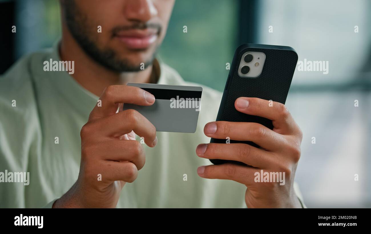 Homme barbu afro-américain à l'intérieur client masculin méconnaissable tenant une carte de crédit et un smartphone assis au bureau à domicile à l'aide d'un paiement mobile facile Banque D'Images