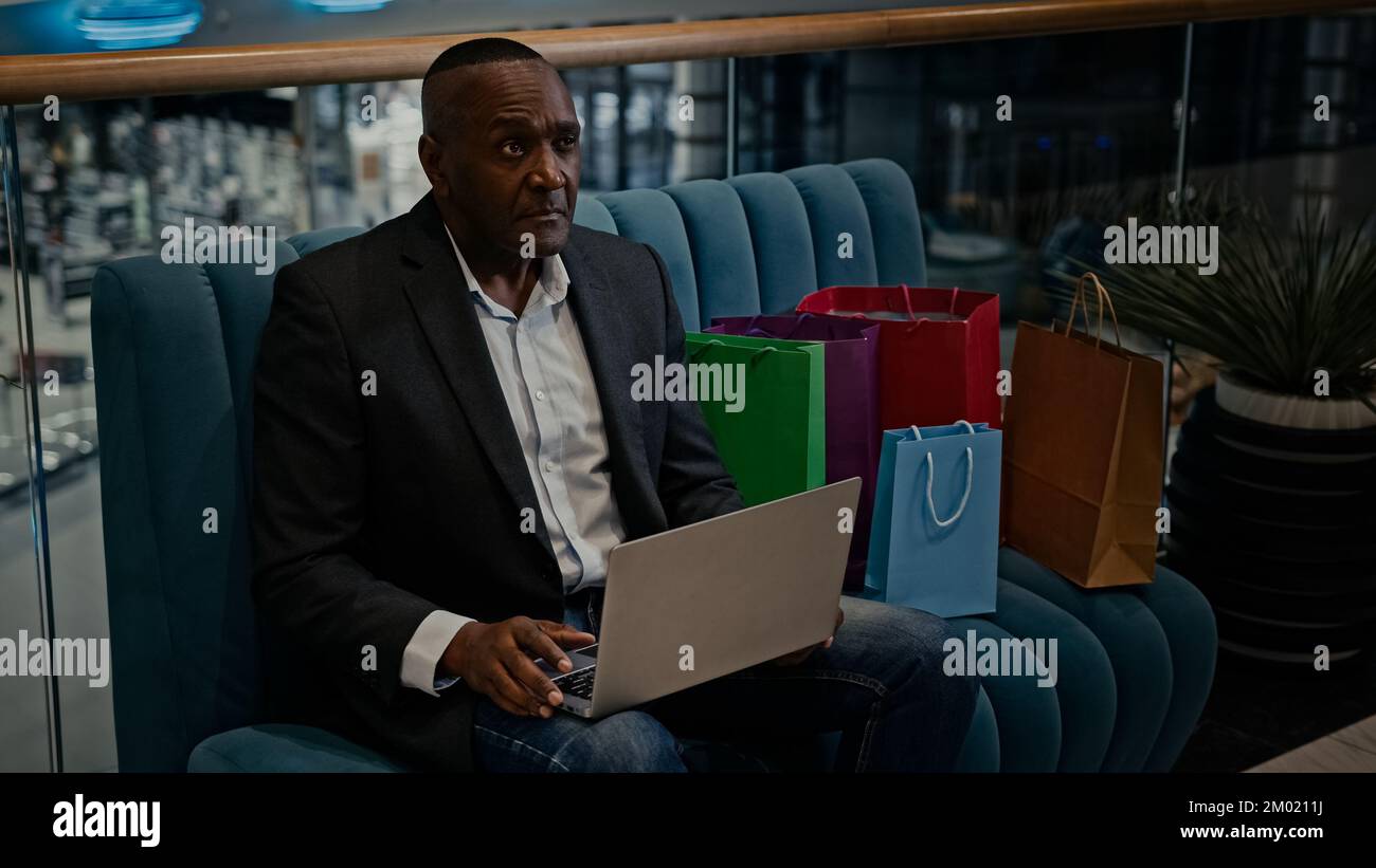 Homme d'affaires américain africain homme d'affaires homme client client dans le centre commercial assis sur un canapé avec des achats de sacs en utilisant l'achat d'ordinateur portable en ligne Banque D'Images