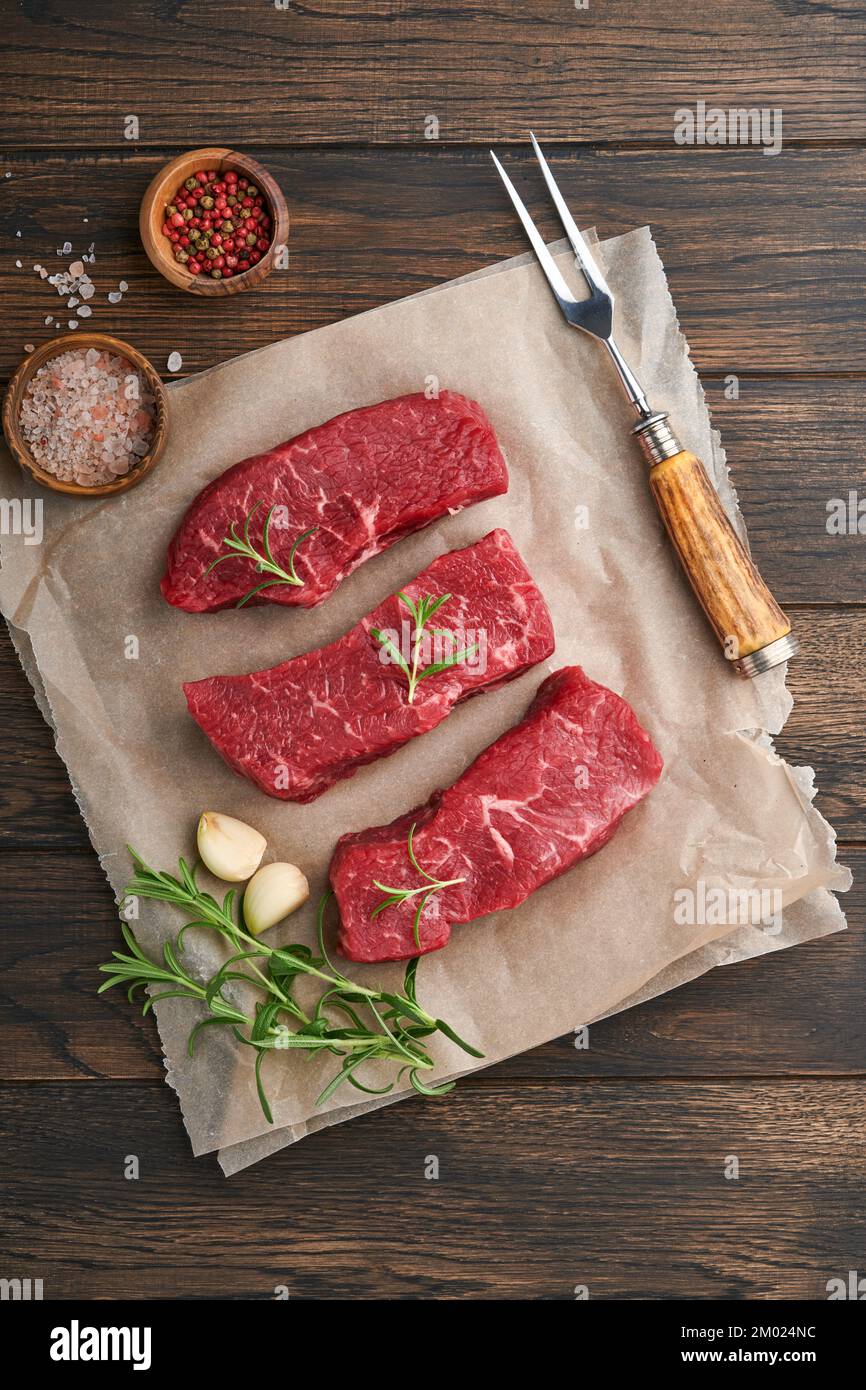 Steaks crus. Sur une planche à bois, placer les steaks sur la lame avec des épices, du romarin, des légumes et des ingrédients pour cuisiner sur un fond de bois ancien. Vue de dessus. Banque D'Images