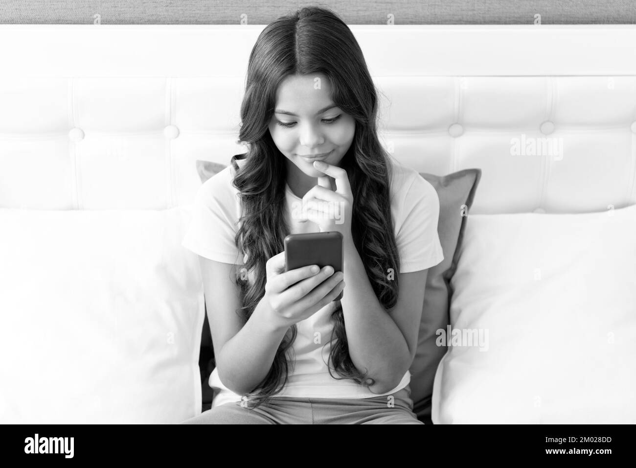 Une fille heureuse sourit en discutant sur un téléphone mobile tout en étant assise sur le lit, en discutant. Banque D'Images