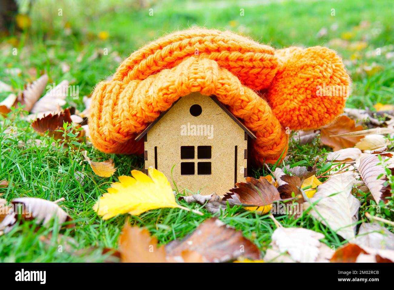 Grosse casquette d'hiver posée sur une figurine de maison en bois placée sur une pelouse verte avec des feuilles d'automne à la campagne. Restez au chaud à la maison sans chauffage. Banque D'Images