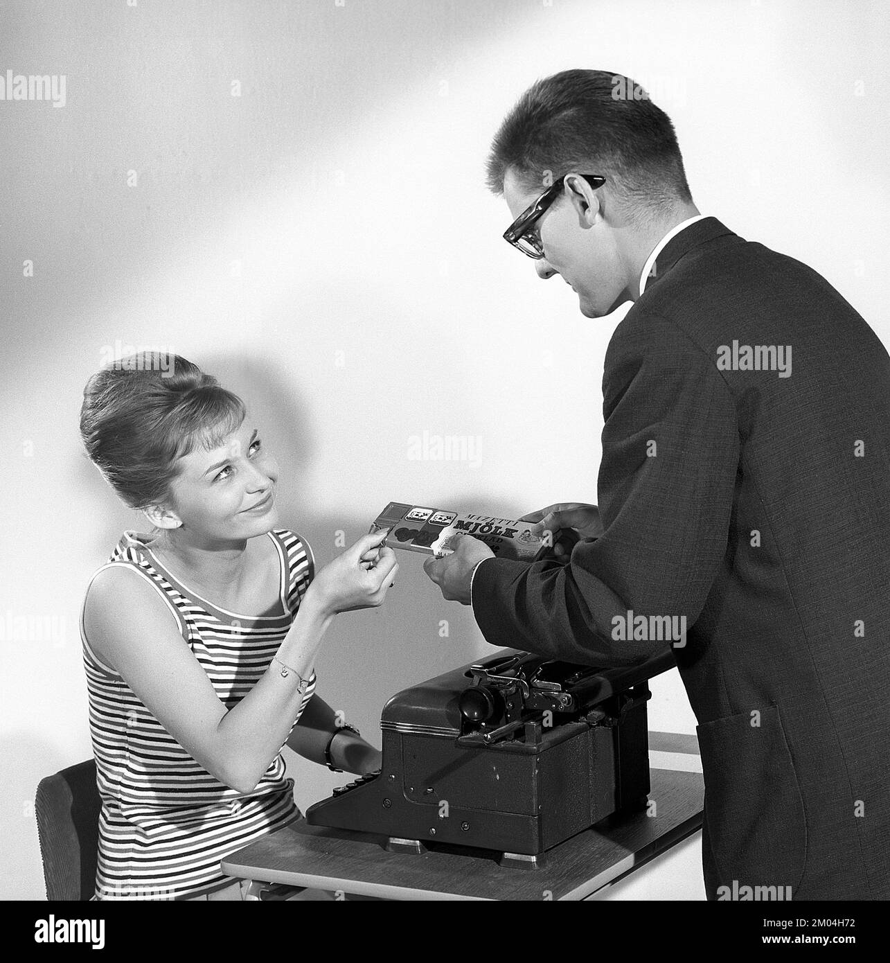 Chocolat dans le 1960s. Un homme offre à une femme un morceau d'une barre de chocolat. Elle est assise par sa machine à écrire. Elle a ses cheveux dans la coiffure de ruche typique, dans laquelle les cheveux longs sont empilés sur le dessus de la tête et donnant une certaine ressemblance à la forme d'une ruche traditionnelle. Suède 1963. Kristoffersson réf. CZ54 Banque D'Images