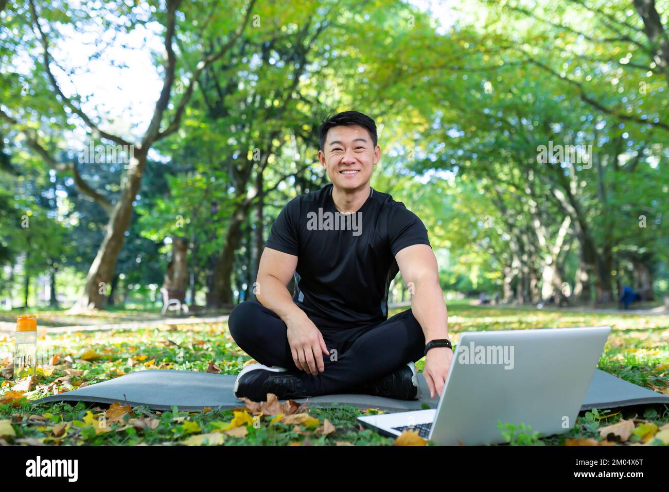 Formation en ligne. Beau homme asiatique, entraîneur sportif assis sur un tapis dans le parc avec un ordinateur portable et conduit l'entraînement à distance avec une caméra vidéo. Il regarde la caméra, sourit. Banque D'Images