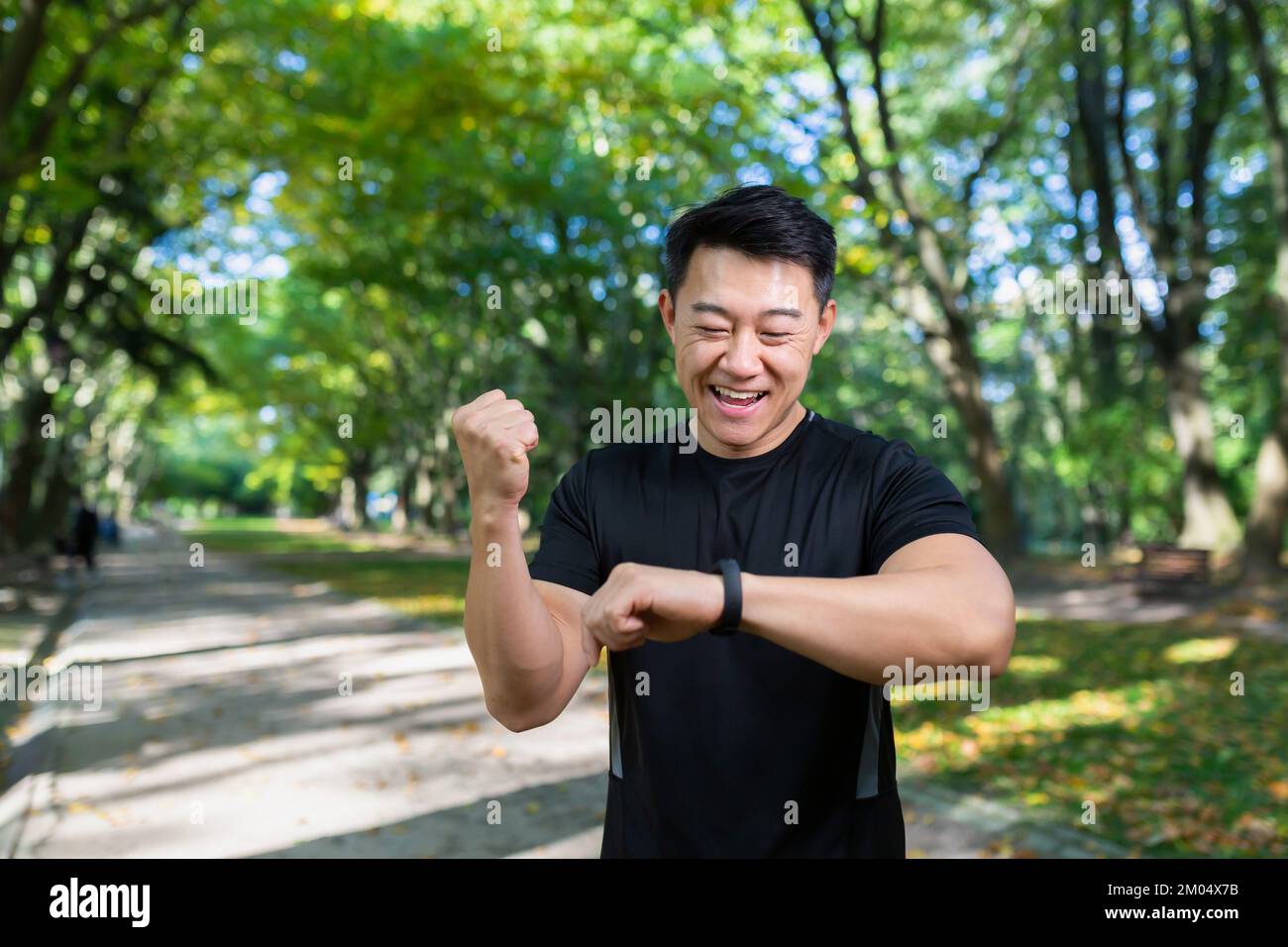 Marathon. Un jeune athlète asiatique se tient dans le parc en vêtements de sport, regarde un bracelet de fitness, une montre intelligente, se réjouit du résultat d'une course, montre un geste gagnant avec sa main. Banque D'Images