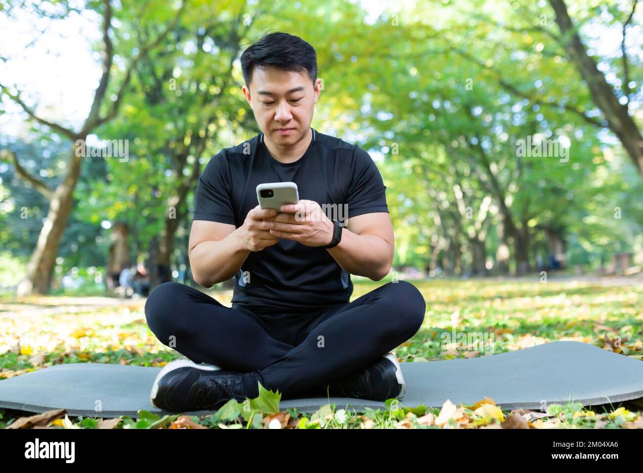 Jeune homme sportif asiatique élégant, assis dans le parc sur un tapis de sport noir, utilise un téléphone portable. Repos après un entraînement actif. Banque D'Images