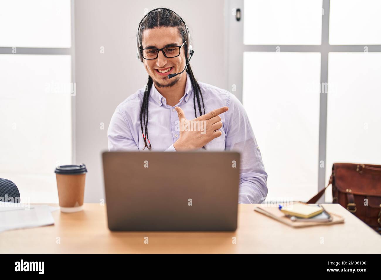 Homme hispanique aux cheveux longs travaillant avec ordinateur portable souriant gai pointant avec la main et le doigt vers le haut sur le côté Banque D'Images
