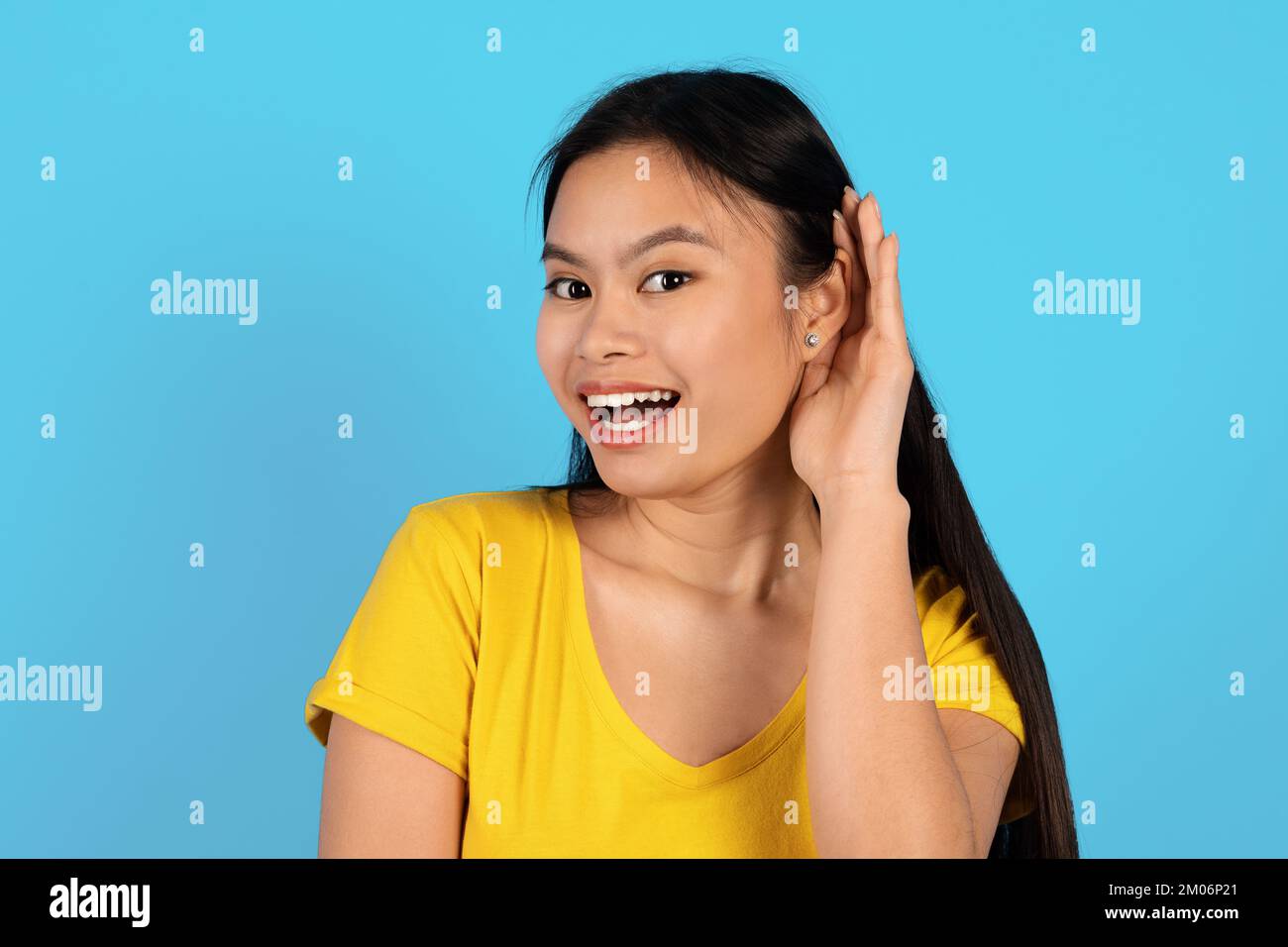 Portrait de la jeune femme coréenne heureuse et inquistive en t-shirt jaune dépasse l'oreille et écoute les nouvelles, regarde l'appareil photo Banque D'Images