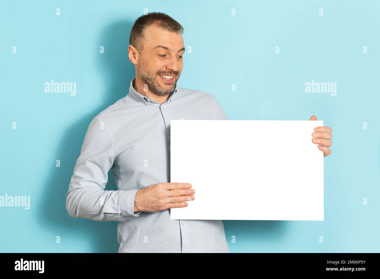 Joyeux homme d'âge moyen montrant un écriteau blanc vierge, posant sur fond bleu de studio, maquette pour logo ou design Banque D'Images