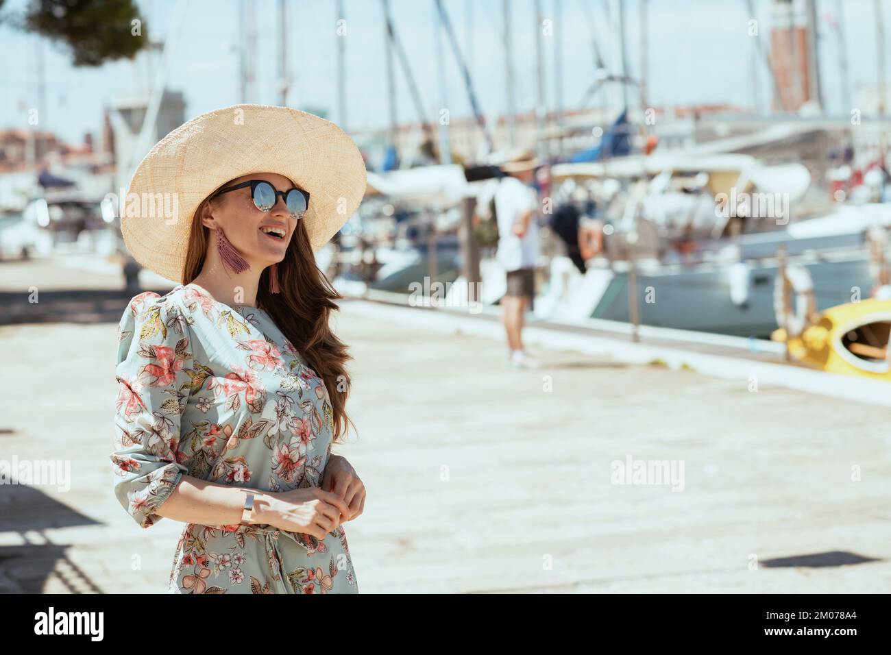 souriante et élégante femme touristique en robe fleurie avec lunettes de  soleil et chapeau sur la jetée Photo Stock - Alamy